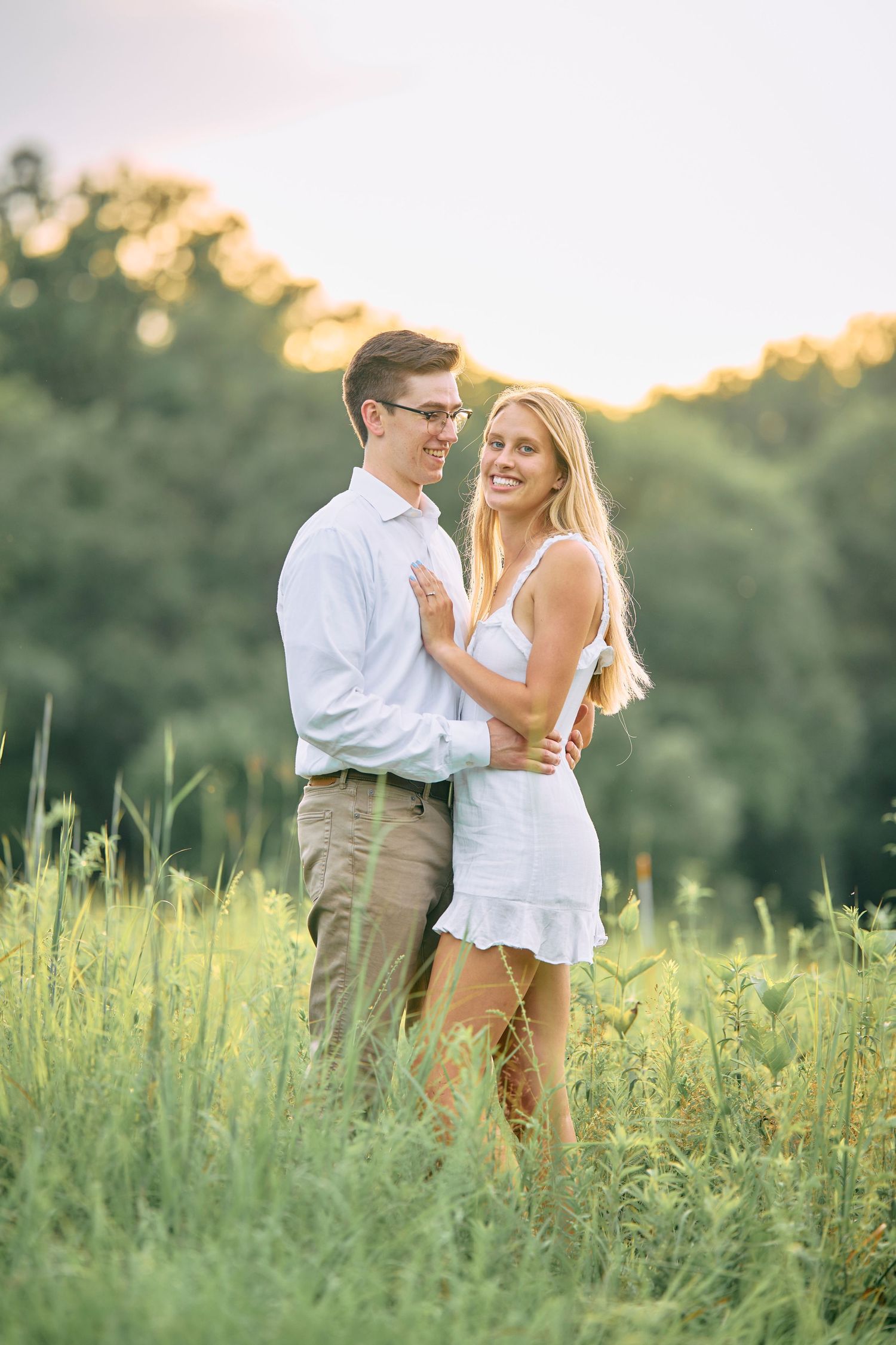 Two people embrace in a romantic moment while standing in a sunlit meadow of tall grass during golden hour.
