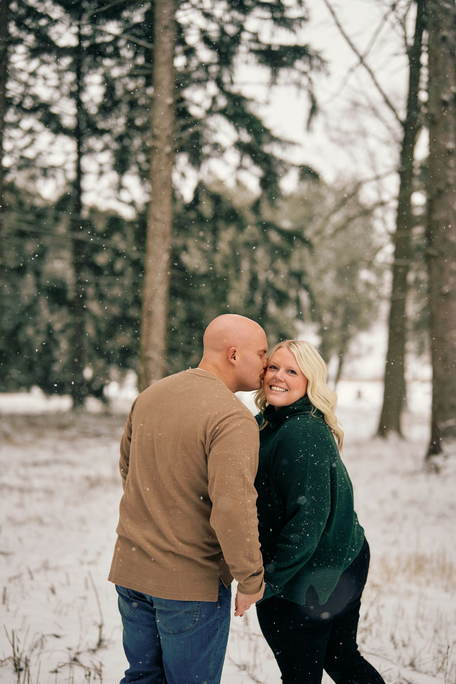 A couple shares an intimate moment while walking together in a snowy forest setting during winter.