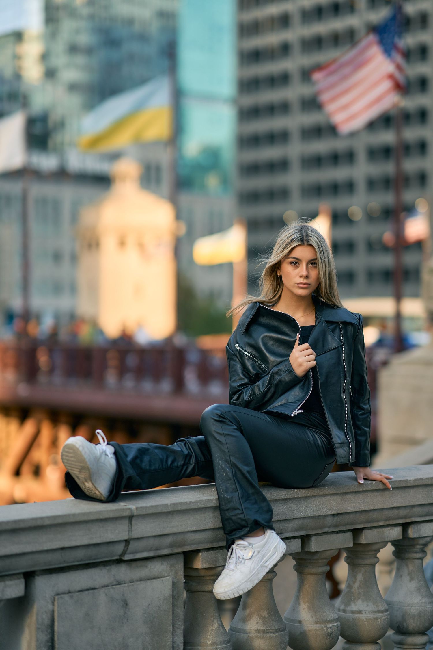 Fashion model in leather jacket and white sneakers poses on stone balustrade with American flag and cityscape in background.