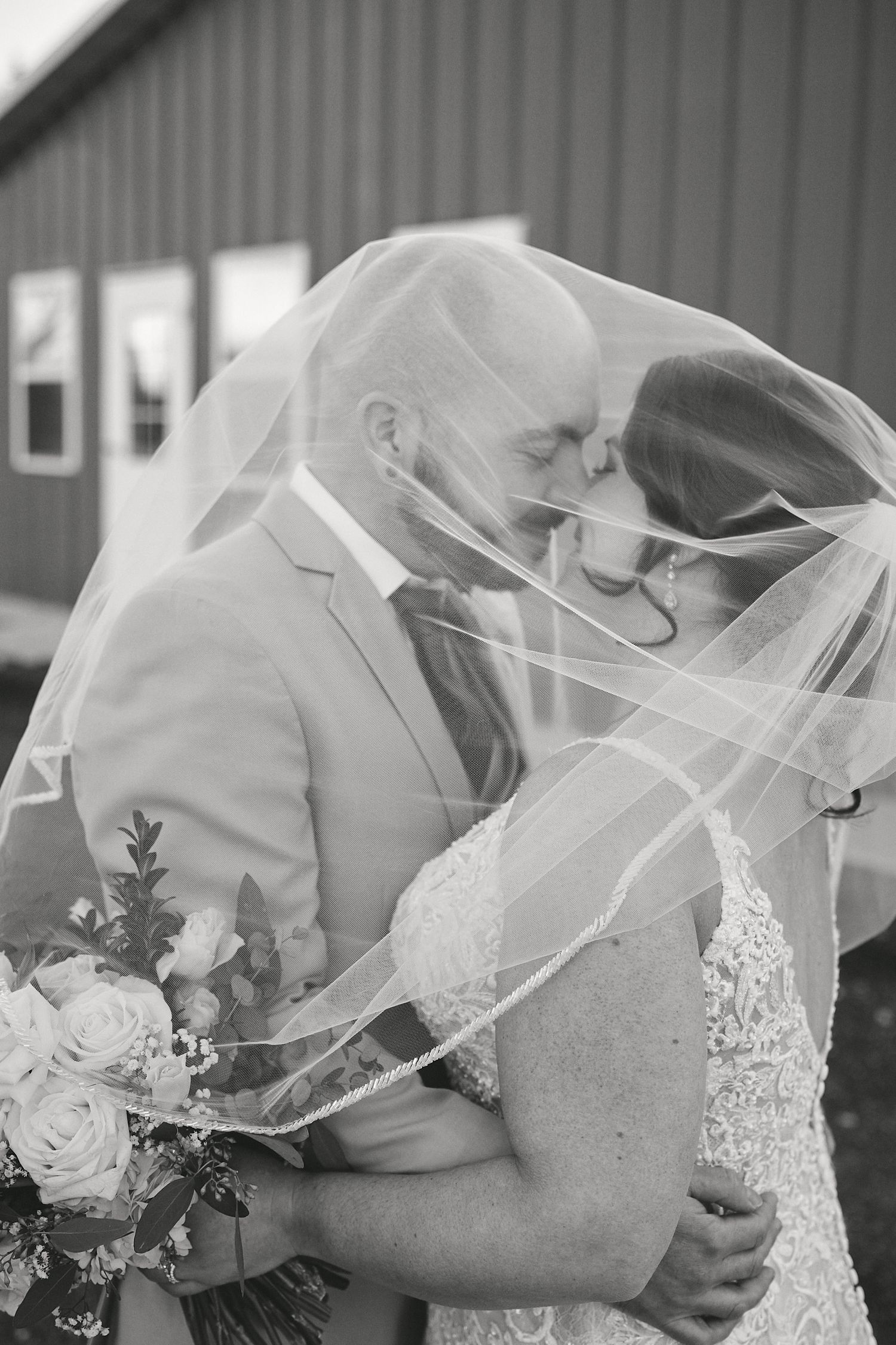 A tender black and white moment between newlyweds sharing a close embrace under a flowing veil beside a rustic building.