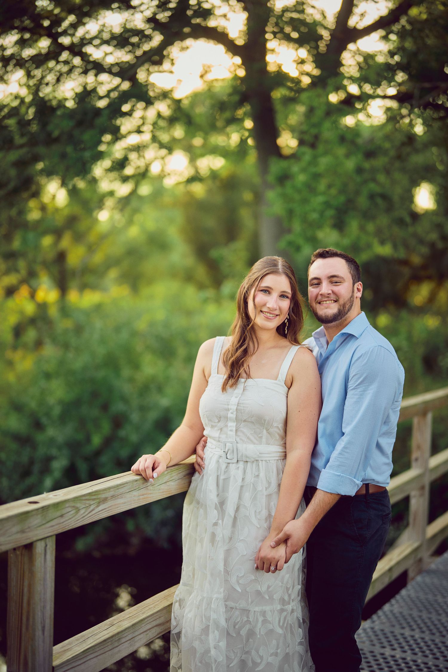 A couple standing on a wooden bridge at sunset surrounded by lush greenery and bokeh lighting effects.