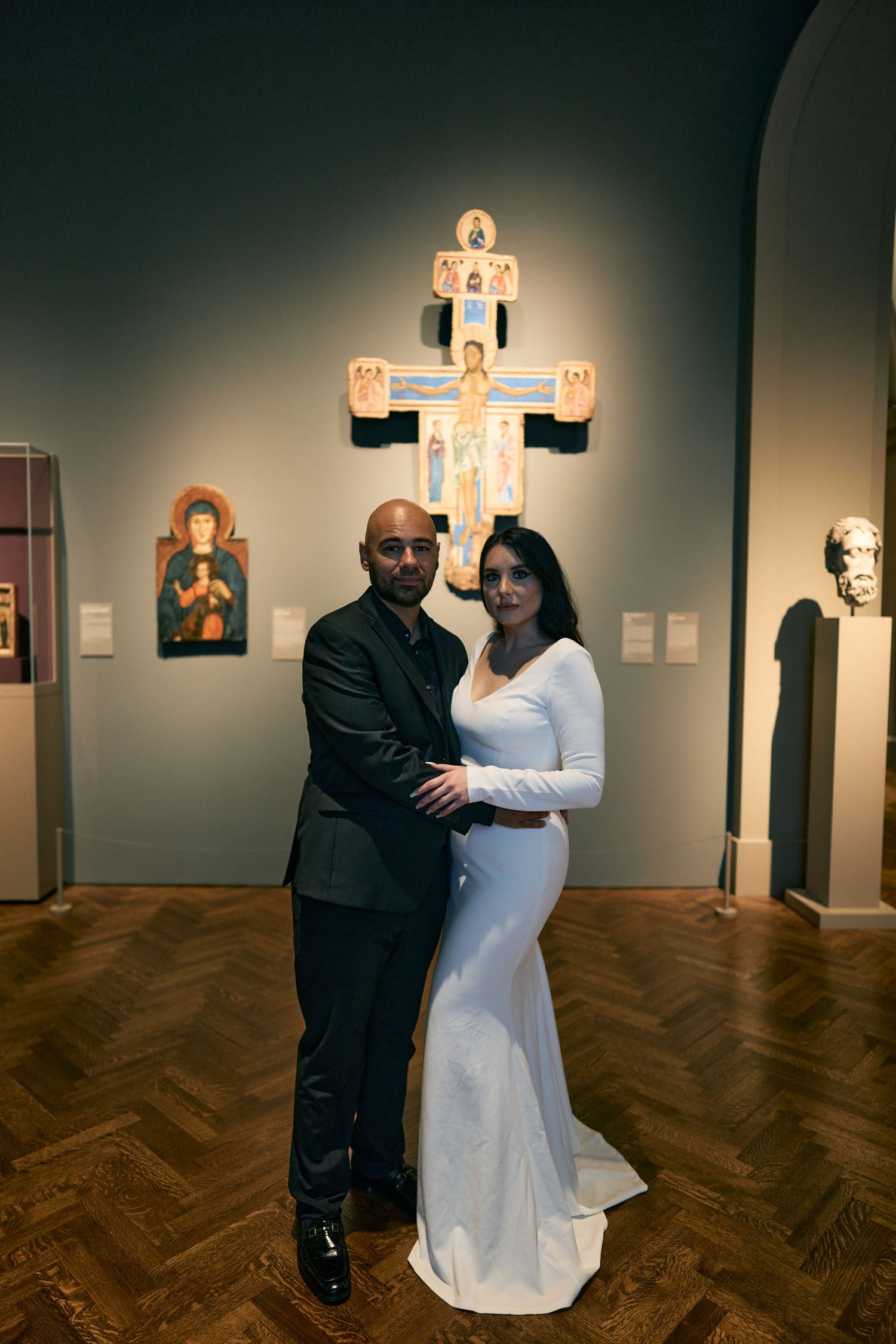 A couple poses together in a museum gallery with formal attire in front of an illuminated religious cross display.