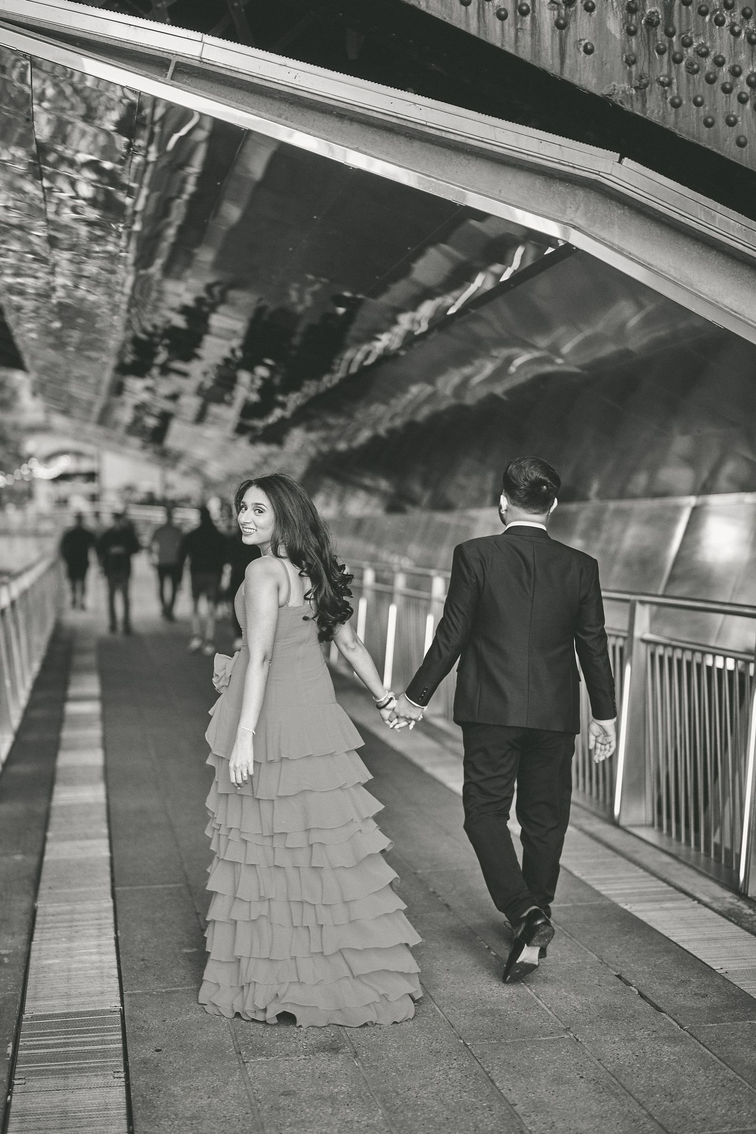A couple in formal attire holding hands while walking through a tunnel with decorative lighting.