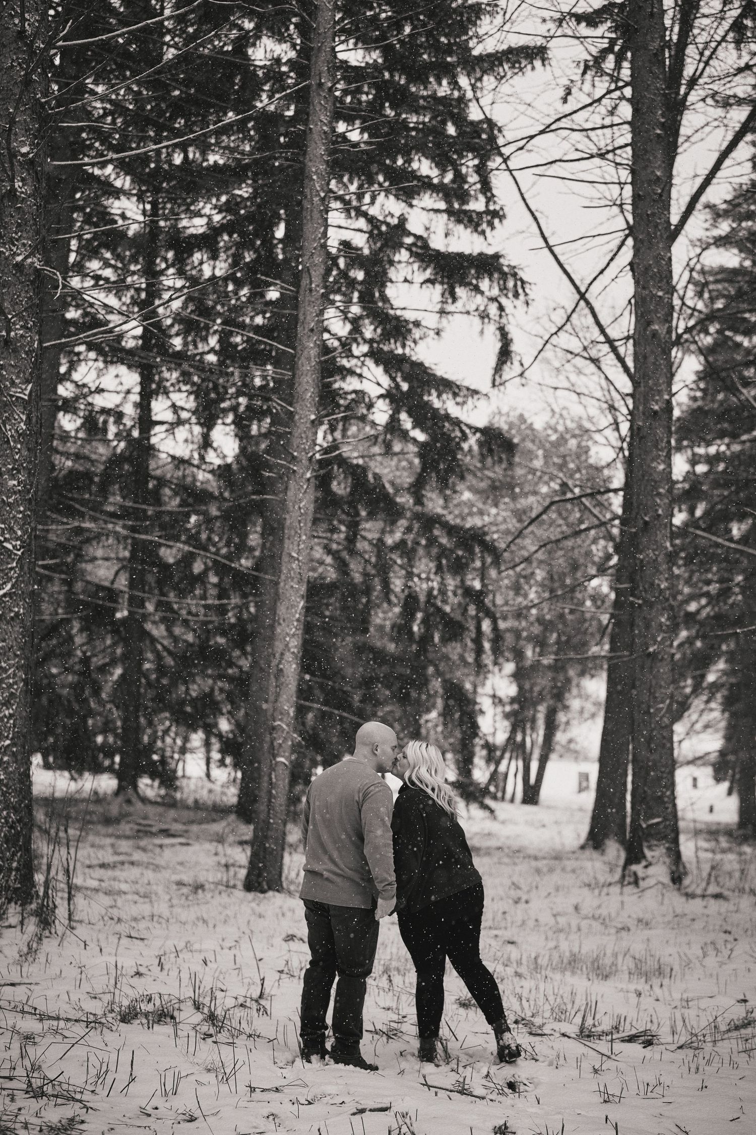 A couple stands together among tall evergreen trees in a snowy winter forest setting, captured in black and white.