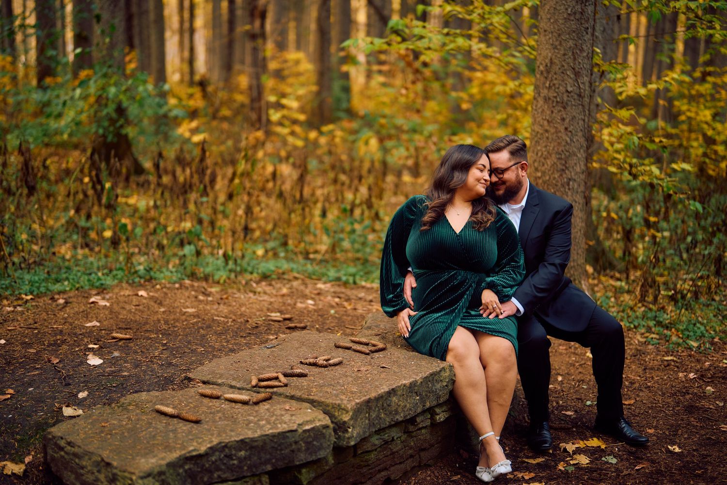 A couple in formal attire sits close together on a stone wall surrounded by autumn foliage in a forest setting.