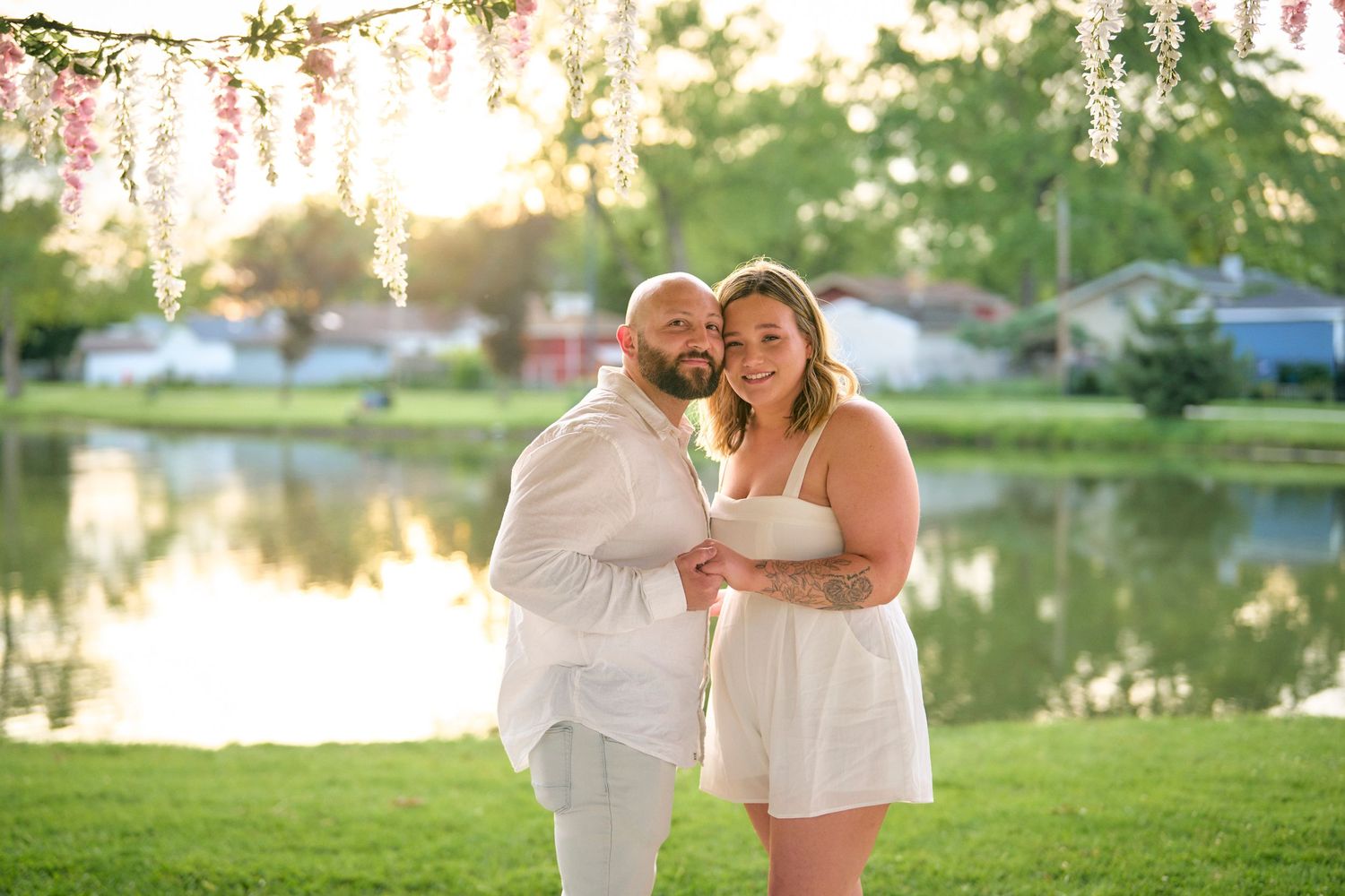 A romantic couple in white outfits poses together by a scenic lake with hanging blossoms at sunset.