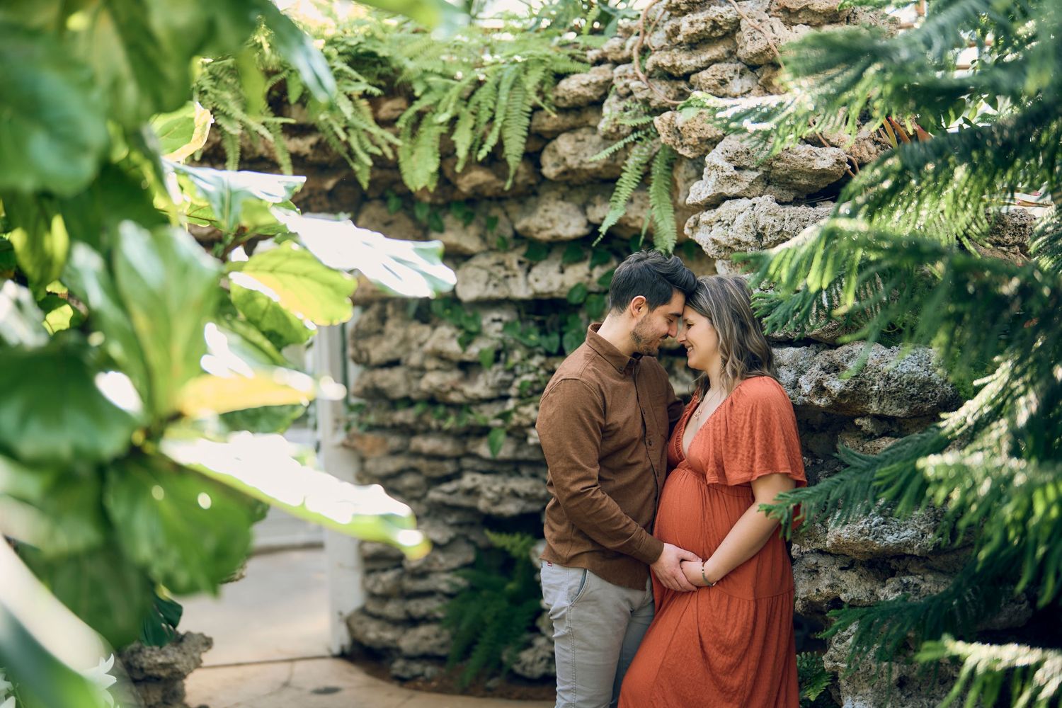 Two people share an intimate moment in the garden beside a rustic stone wall surrounded by lush greenery.
