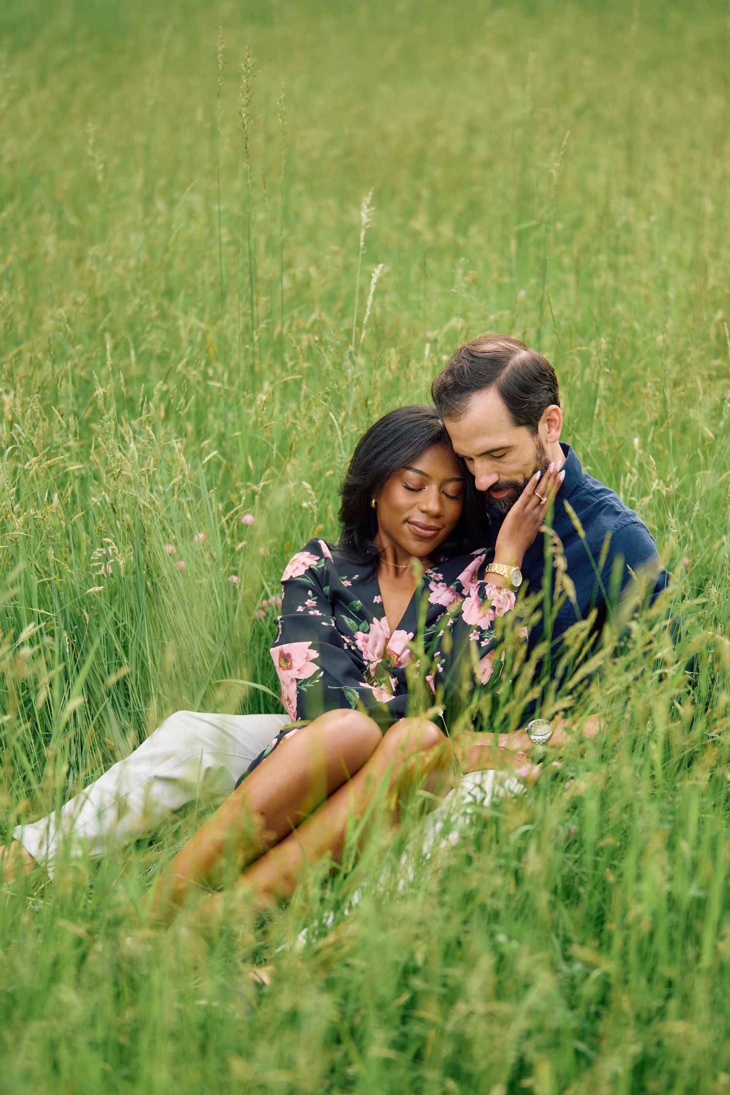 A loving couple embraces while sitting together in tall grass on a sunny summer day in a peaceful meadow.