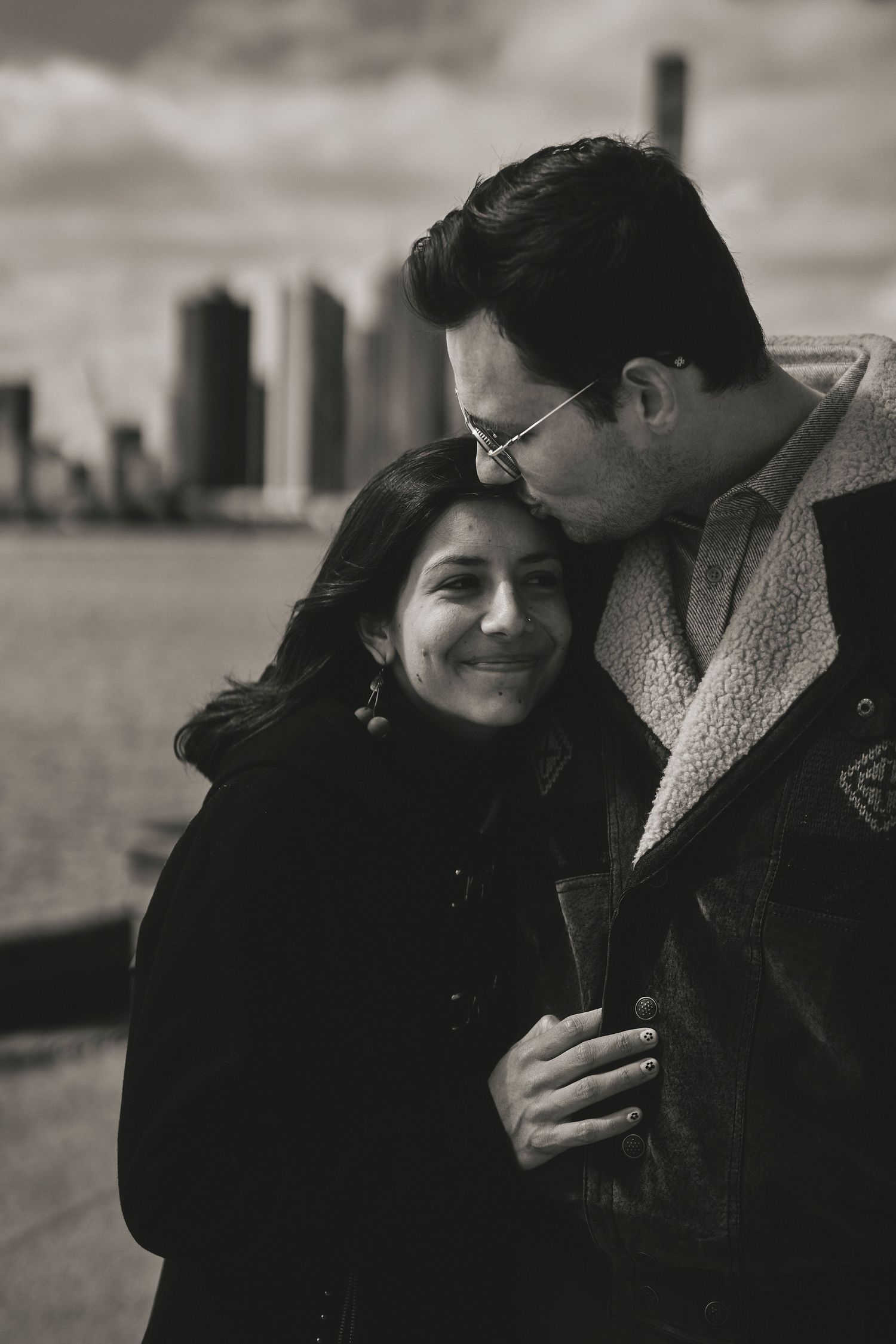 A romantic black and white portrait of a couple embracing by the waterfront with a city skyline in the background.