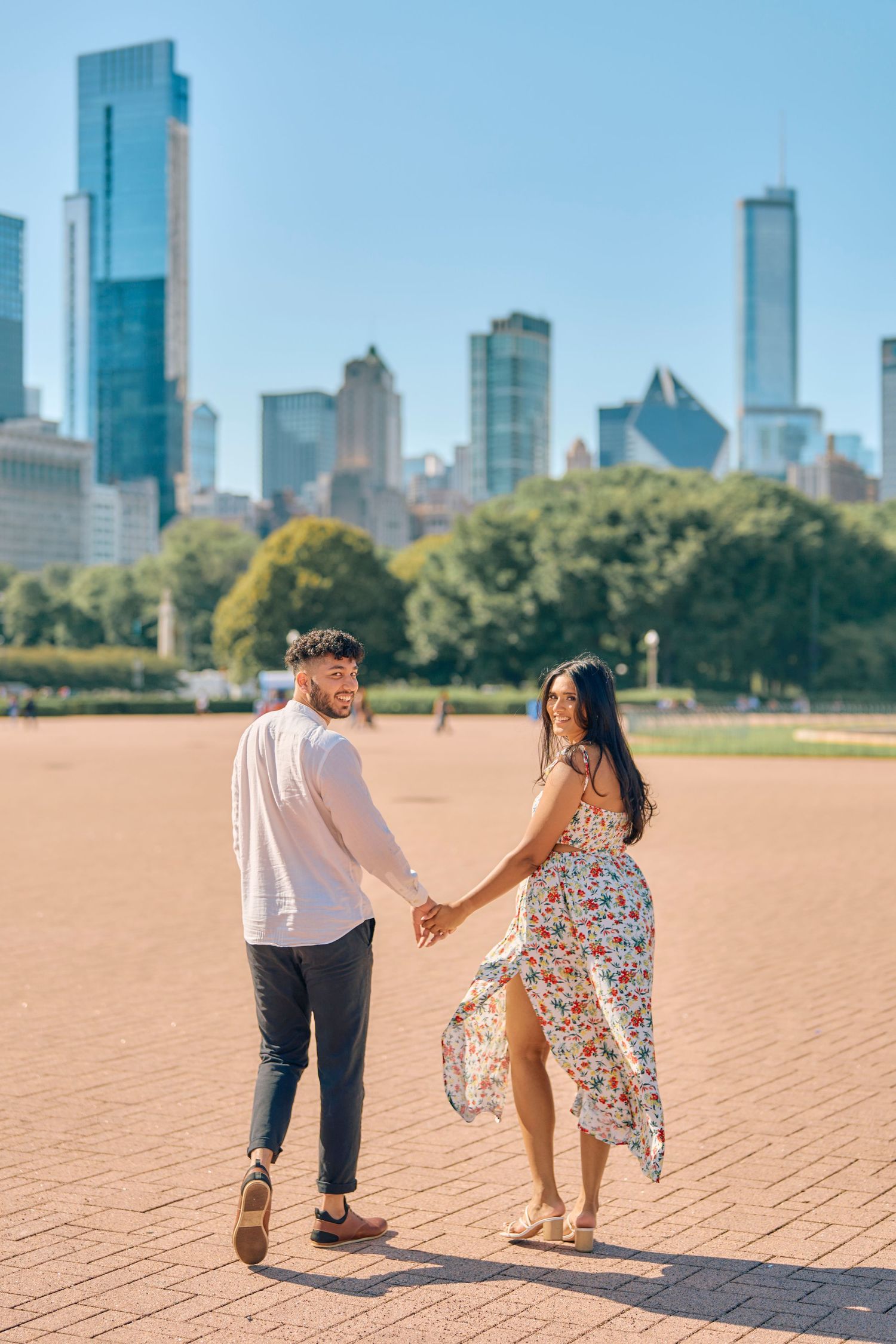 A couple holds hands while walking through Grant Park with the Chicago skyline towering in the background.