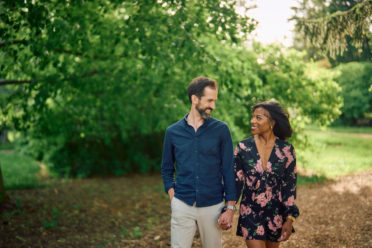 A couple walks together and shares a laugh while strolling down a tree-lined path in a lush garden setting.