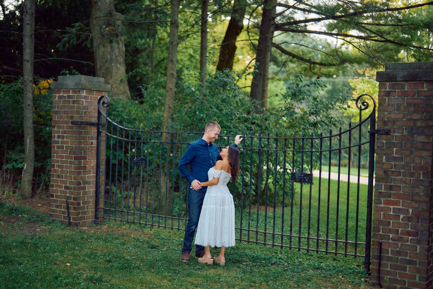 A romantic couple shares a tender moment by ornate iron gates and brick pillars in a lush garden setting at sunset.