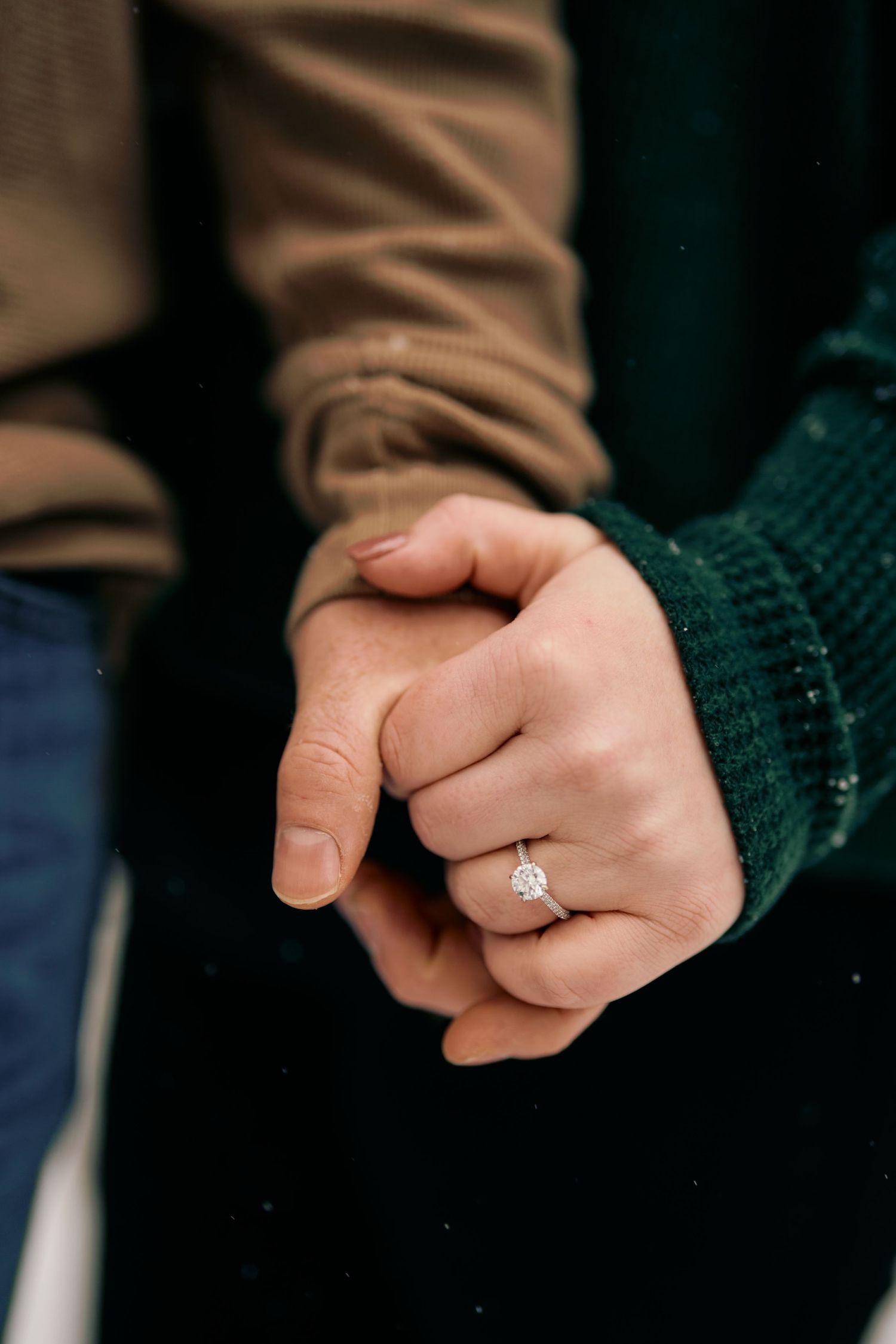 Close-up of two hands holding each other, with one wearing a diamond engagement ring and a green knit sleeve.