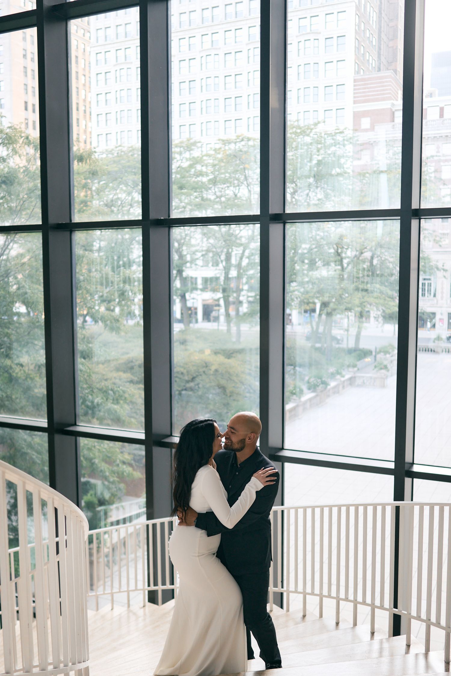 A couple embraces on an elegant indoor staircase with floor-to-ceiling windows overlooking city trees and buildings.