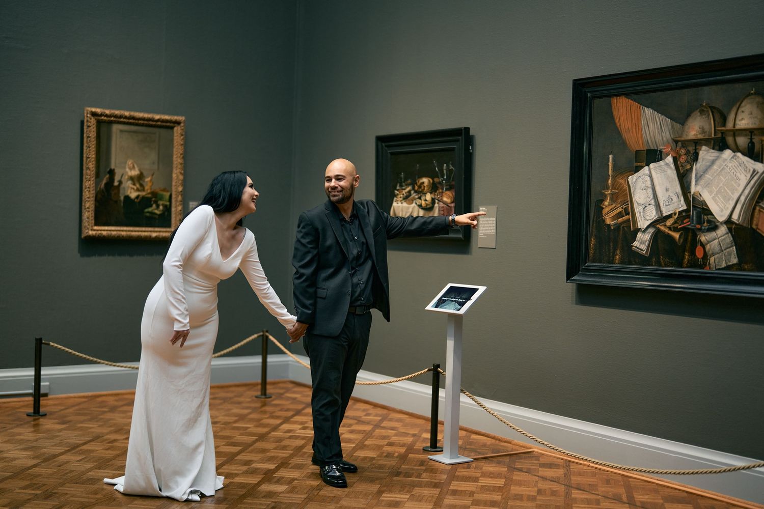 A couple in formal attire views artwork together in an art gallery with green walls and framed paintings.