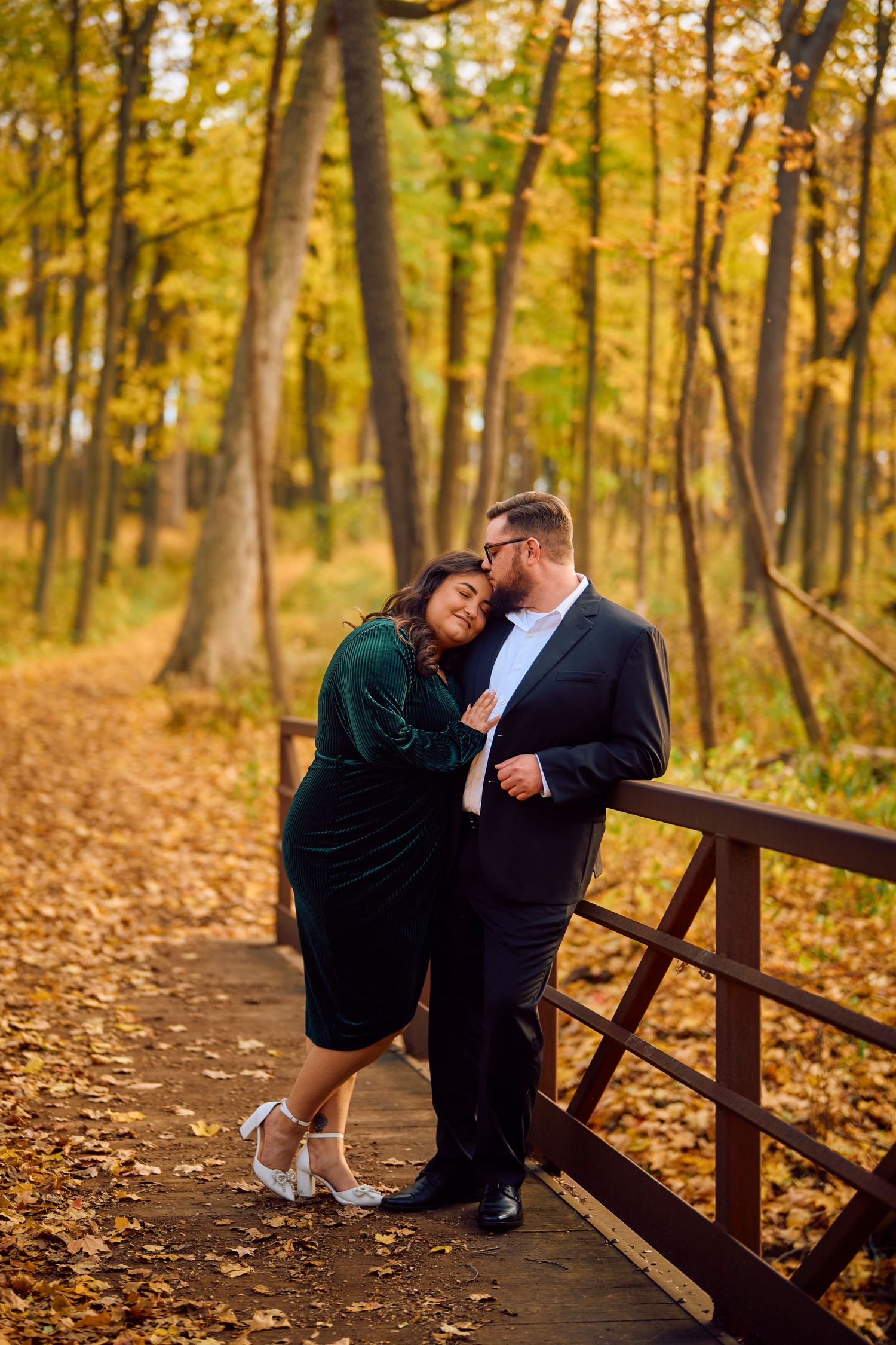 A couple embraces on a wooden bridge surrounded by autumn trees with golden leaves falling in a forest setting.