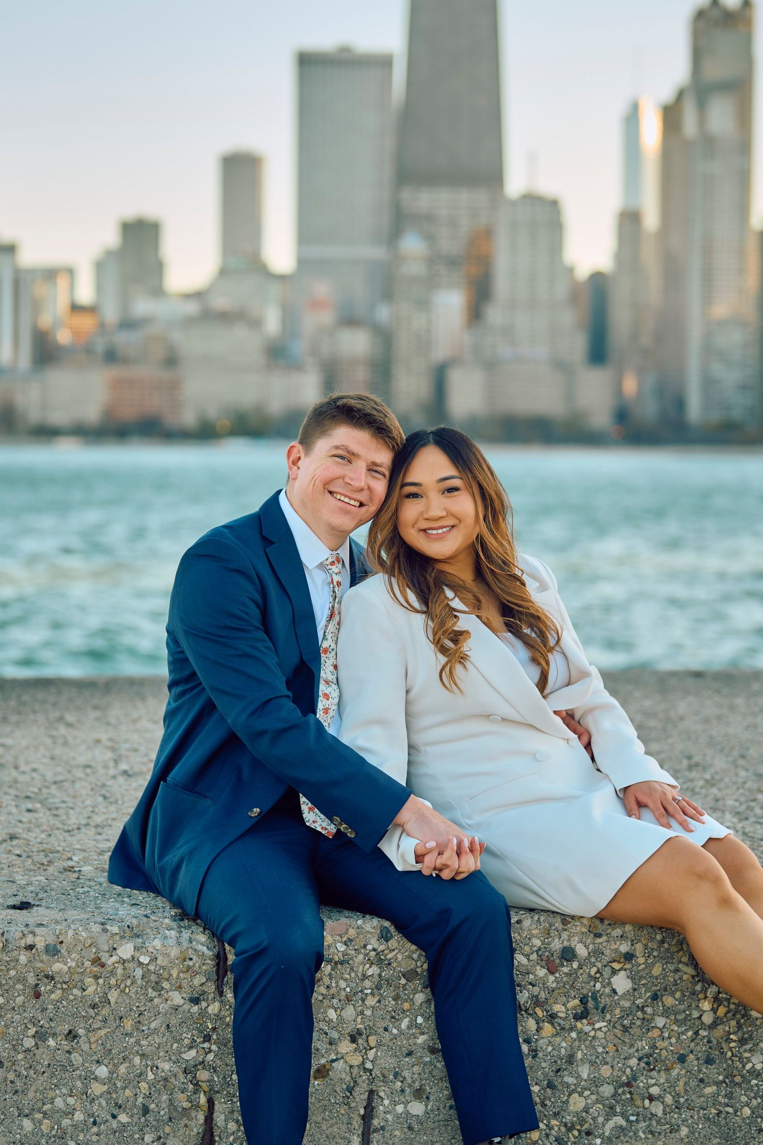 A couple sits together on the shoreline with Chicago's iconic skyline visible across Lake Michigan at sunset.