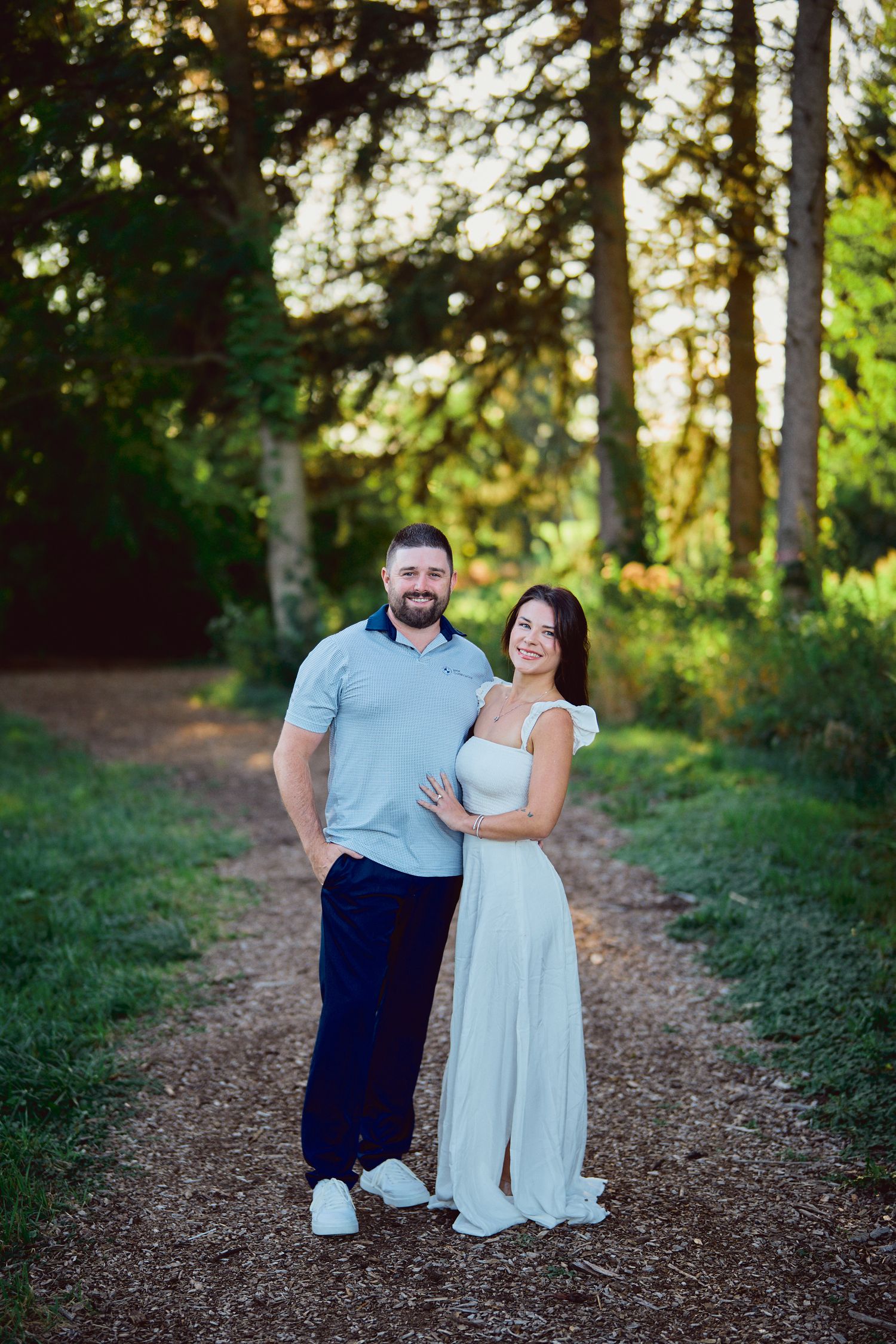 A couple stands together on a forest trail at sunset, dressed in a blue polo and flowing white dress.