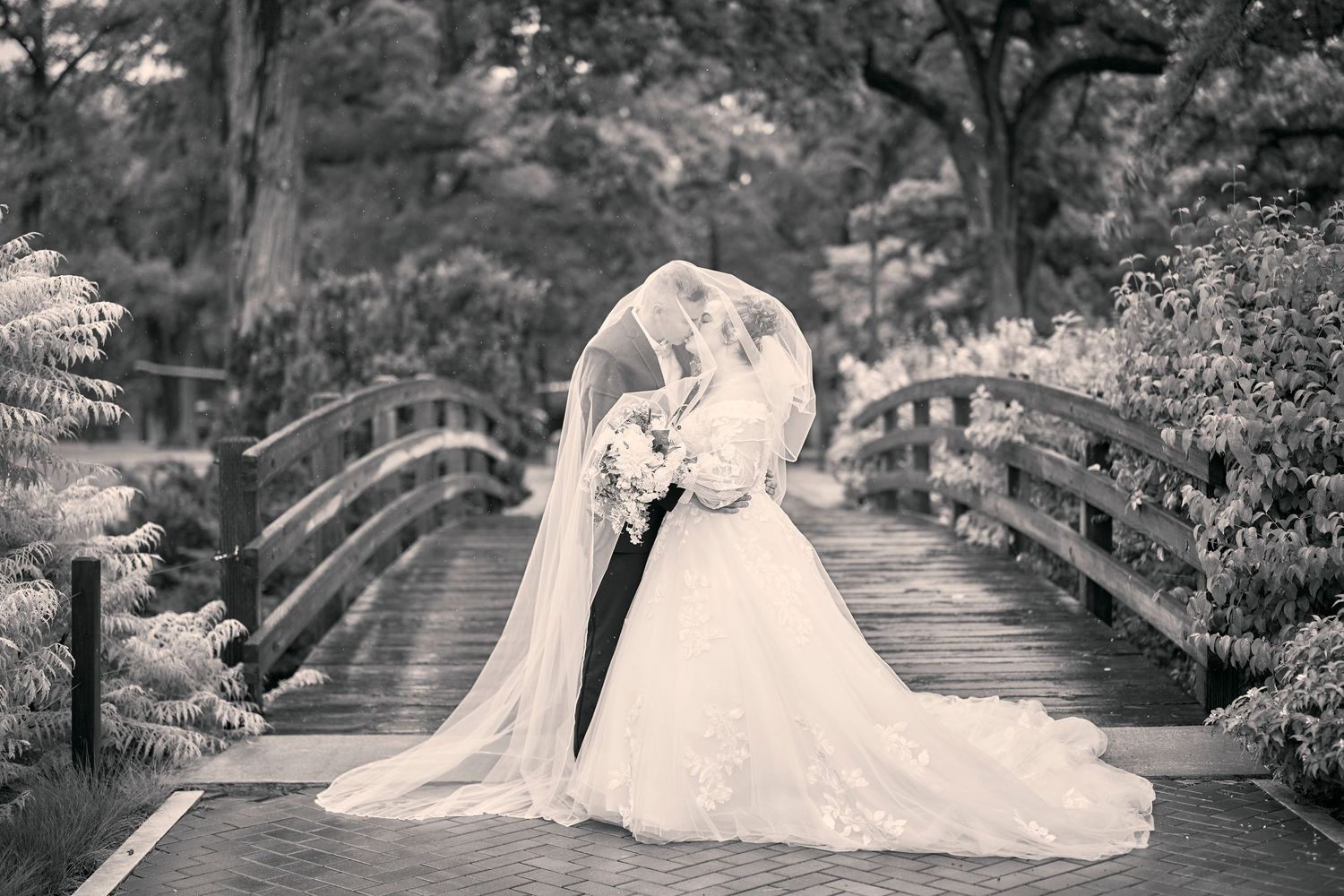 Elegant bride and groom share an intimate moment on a wooden bridge surrounded by lush gardens.