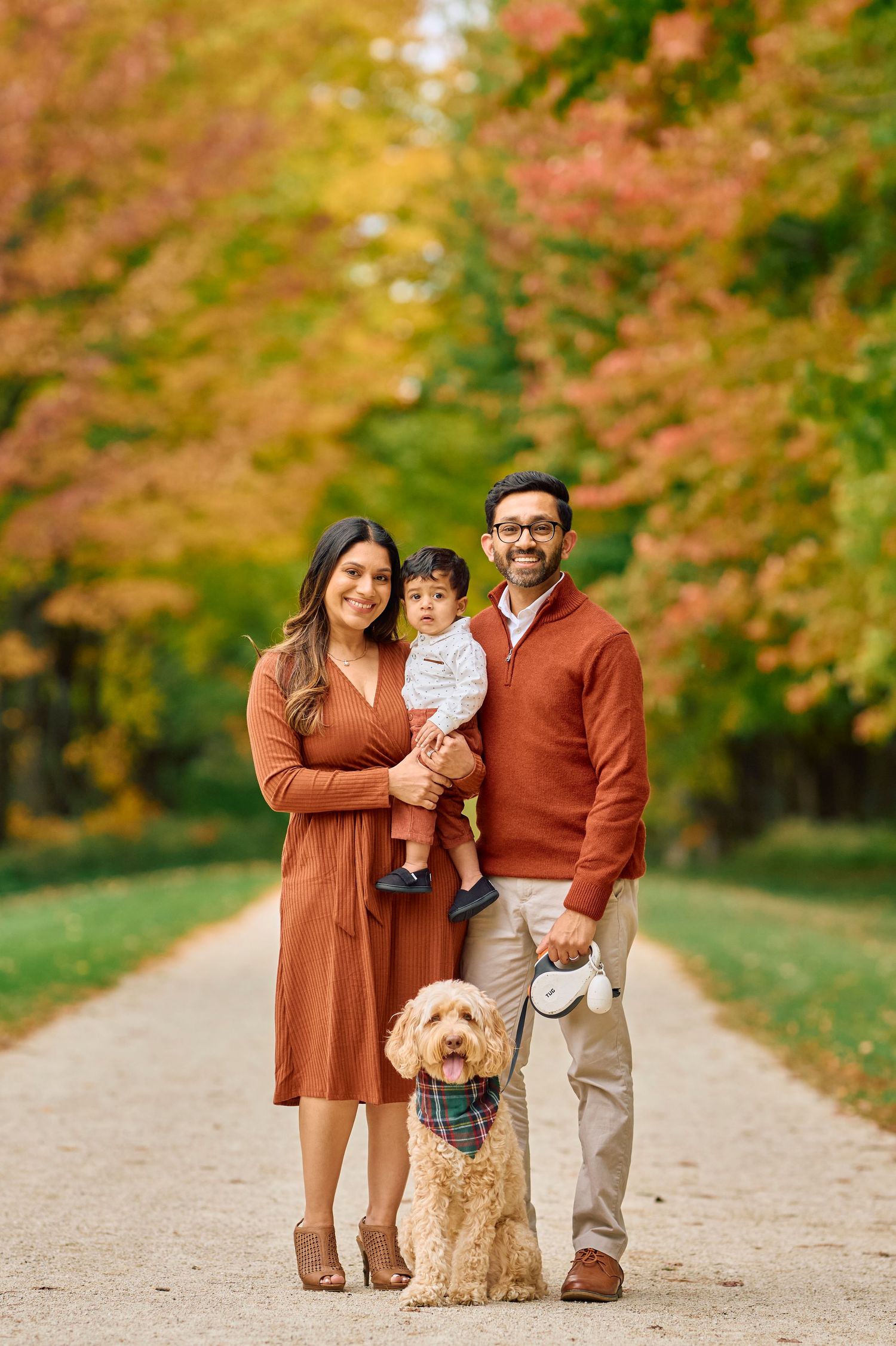 Family portrait on an autumn path with their goldendoodle dog, surrounded by colorful fall foliage in a park.
