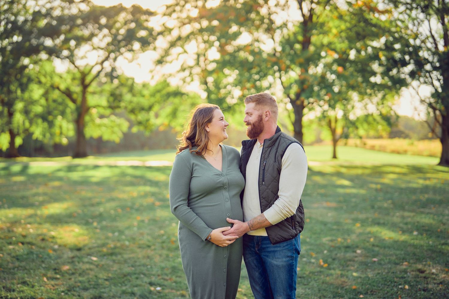 A loving couple shares a tender moment in a sunlit park during an autumn maternity photoshoot.