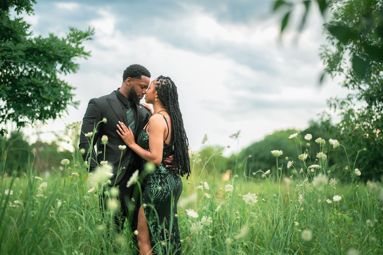 A romantic moment shared in a lush green meadow with wildflowers under a cloudy summer sky.