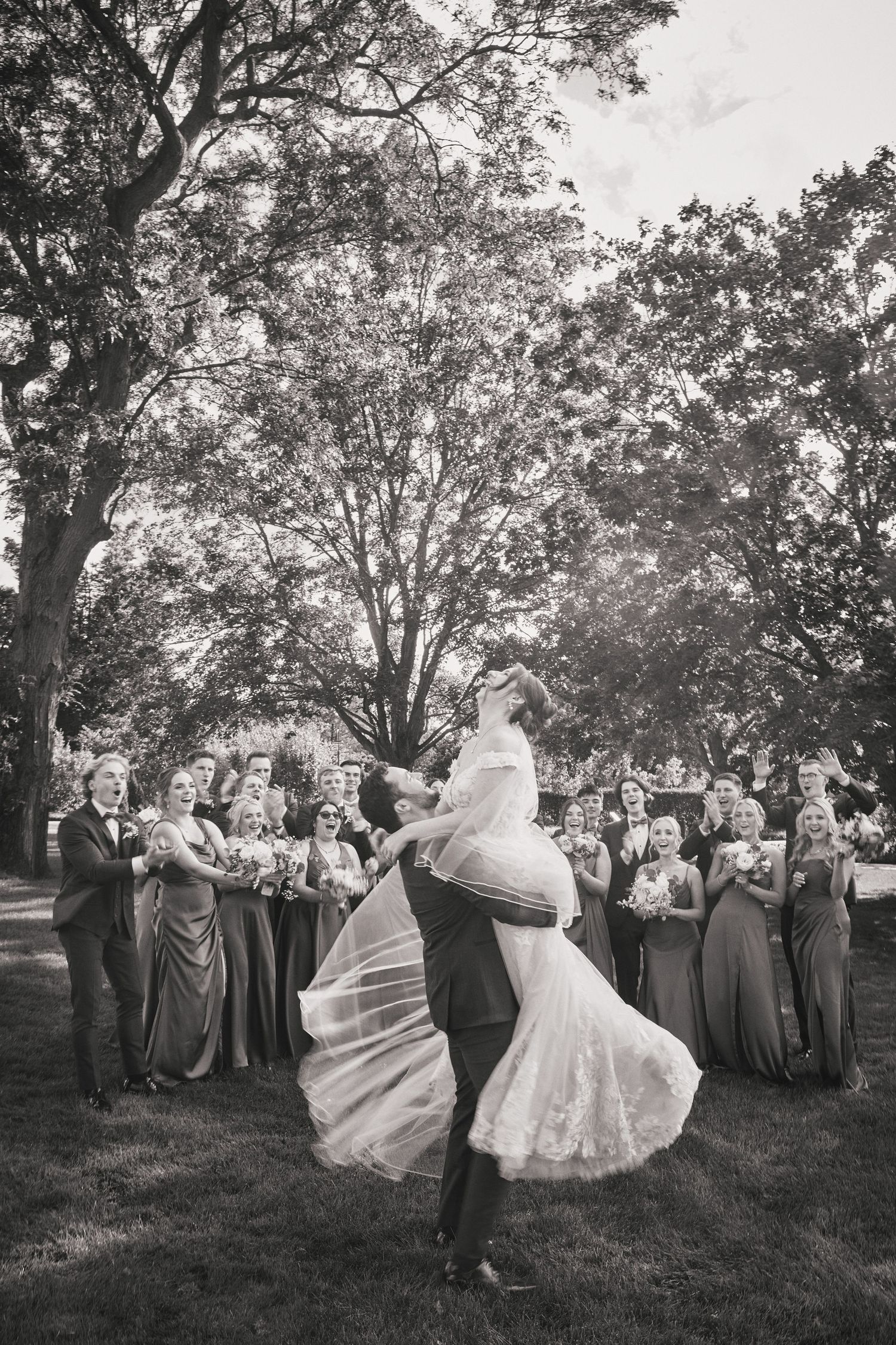 A couple shares their first dance outdoors surrounded by wedding party members in gray dresses under tall trees.
