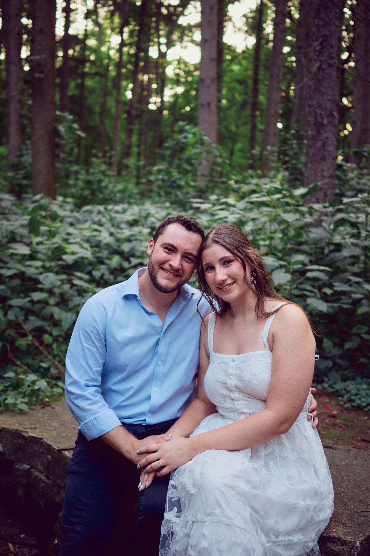 Couple sits together on log in forest setting with lush green foliage in background.