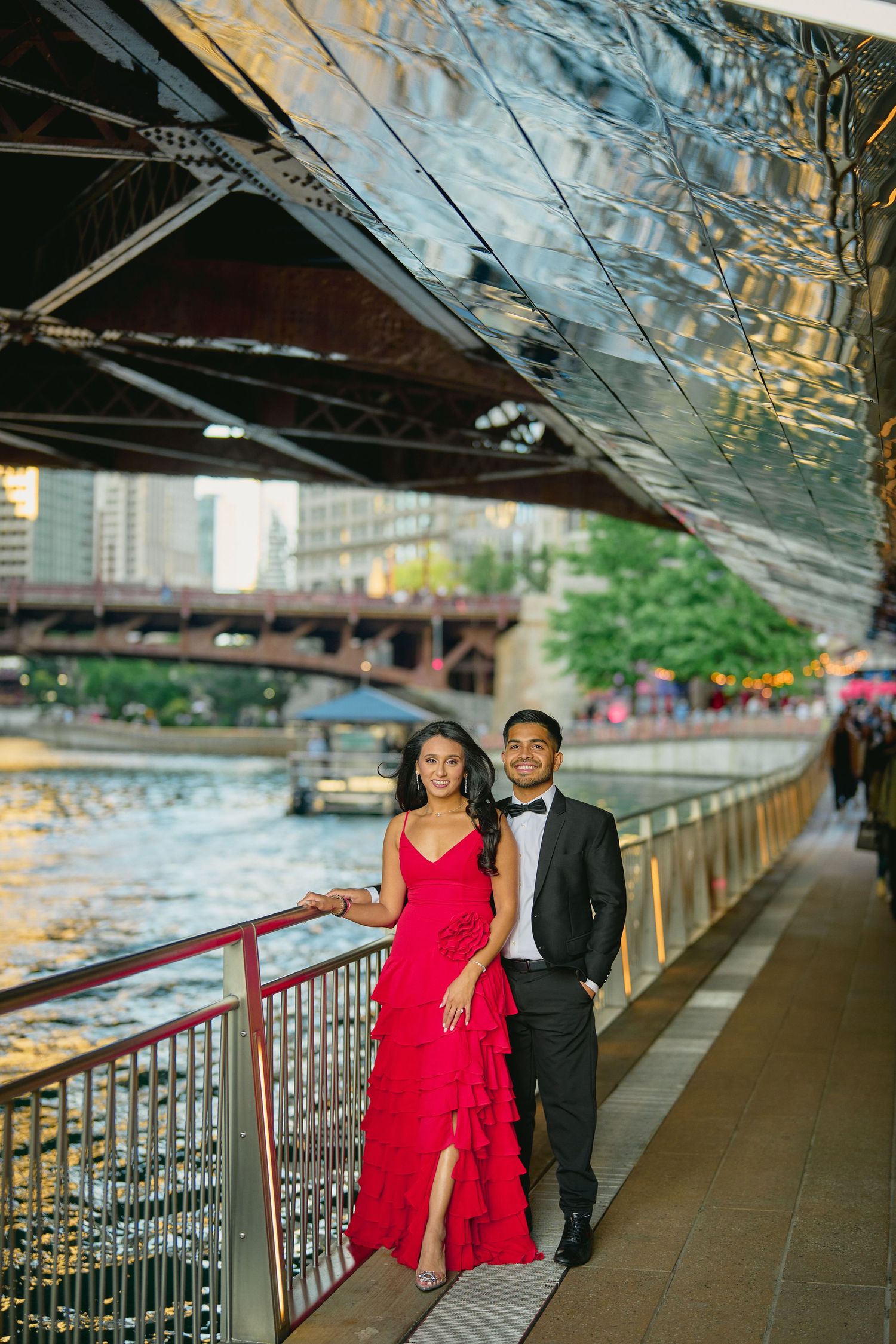 Couple in formal attire poses along Chicago Riverwalk under bridge with red dress and tuxedo during sunset.