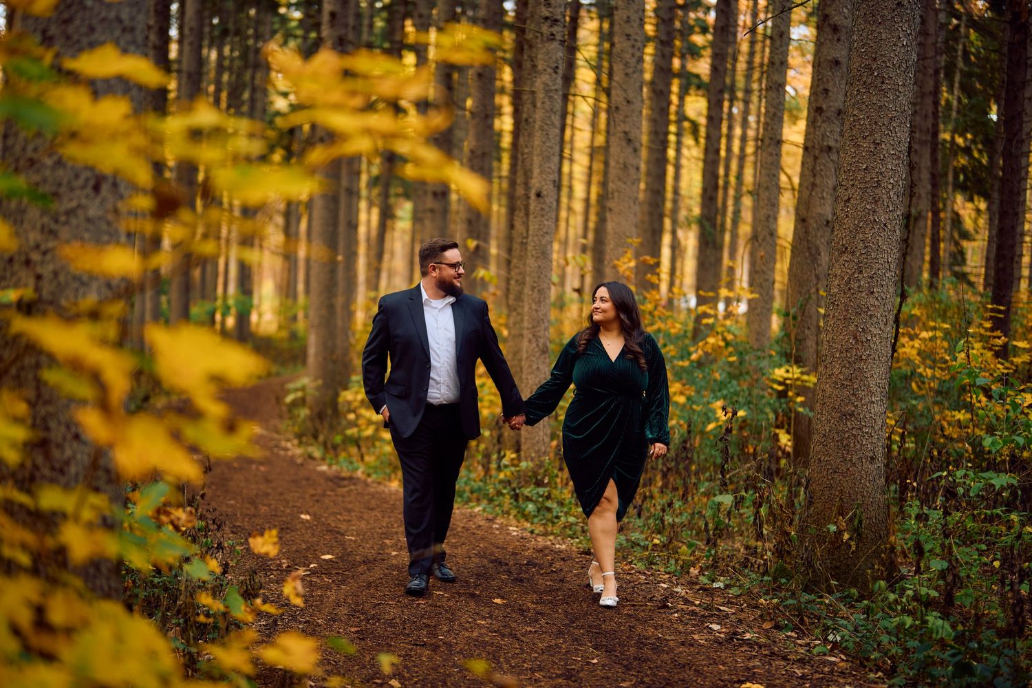 A couple walks hand in hand along a forest trail surrounded by golden autumn foliage and tall pine trees.