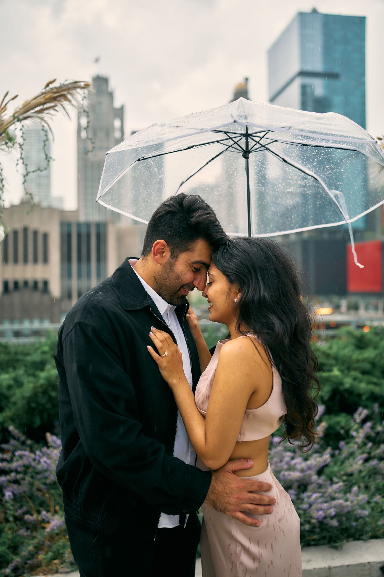Romantic couple sharing an intimate moment under a clear umbrella in an urban garden with city buildings in background.