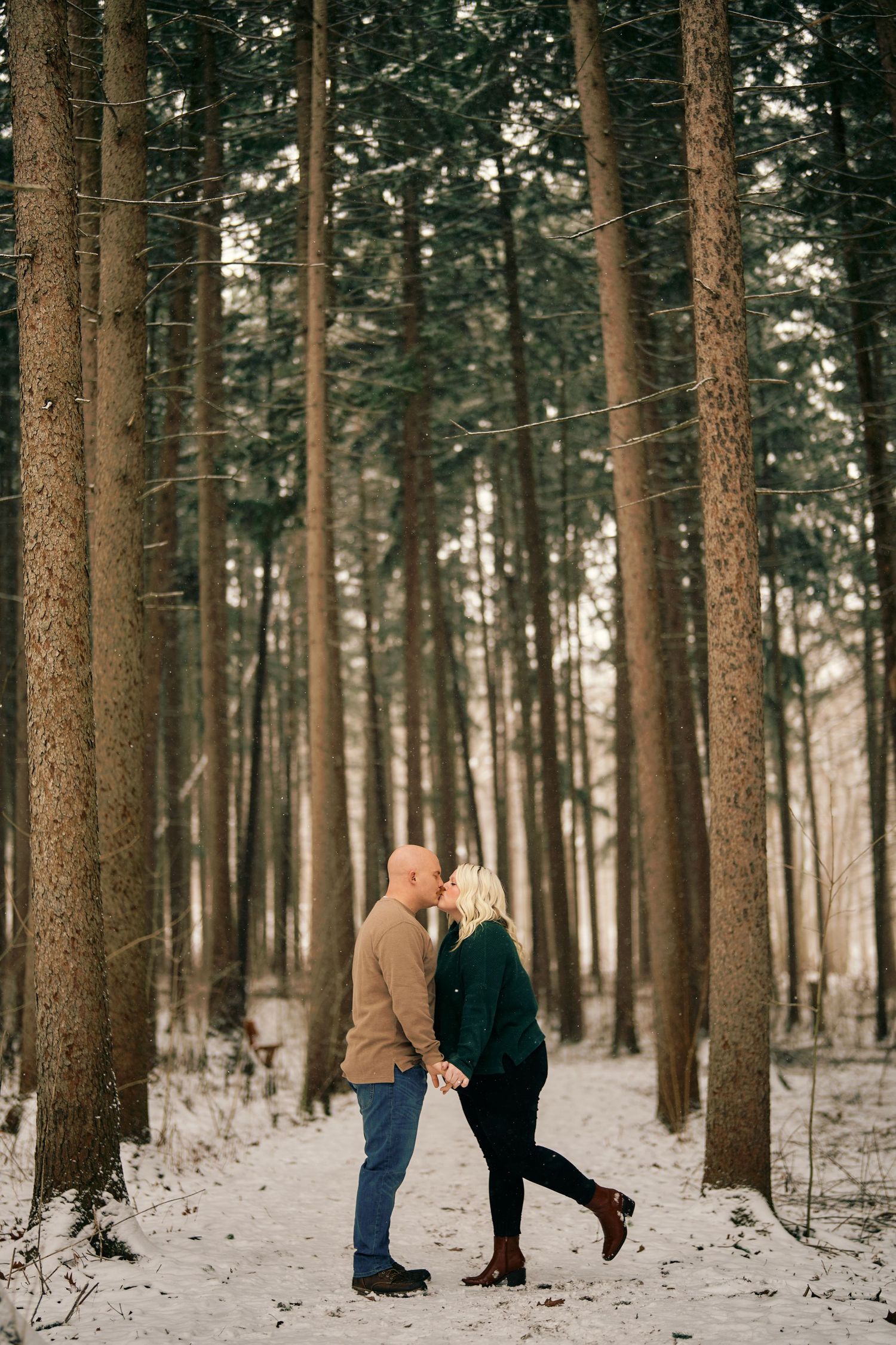 A couple stands close together in a snowy pine forest during winter.