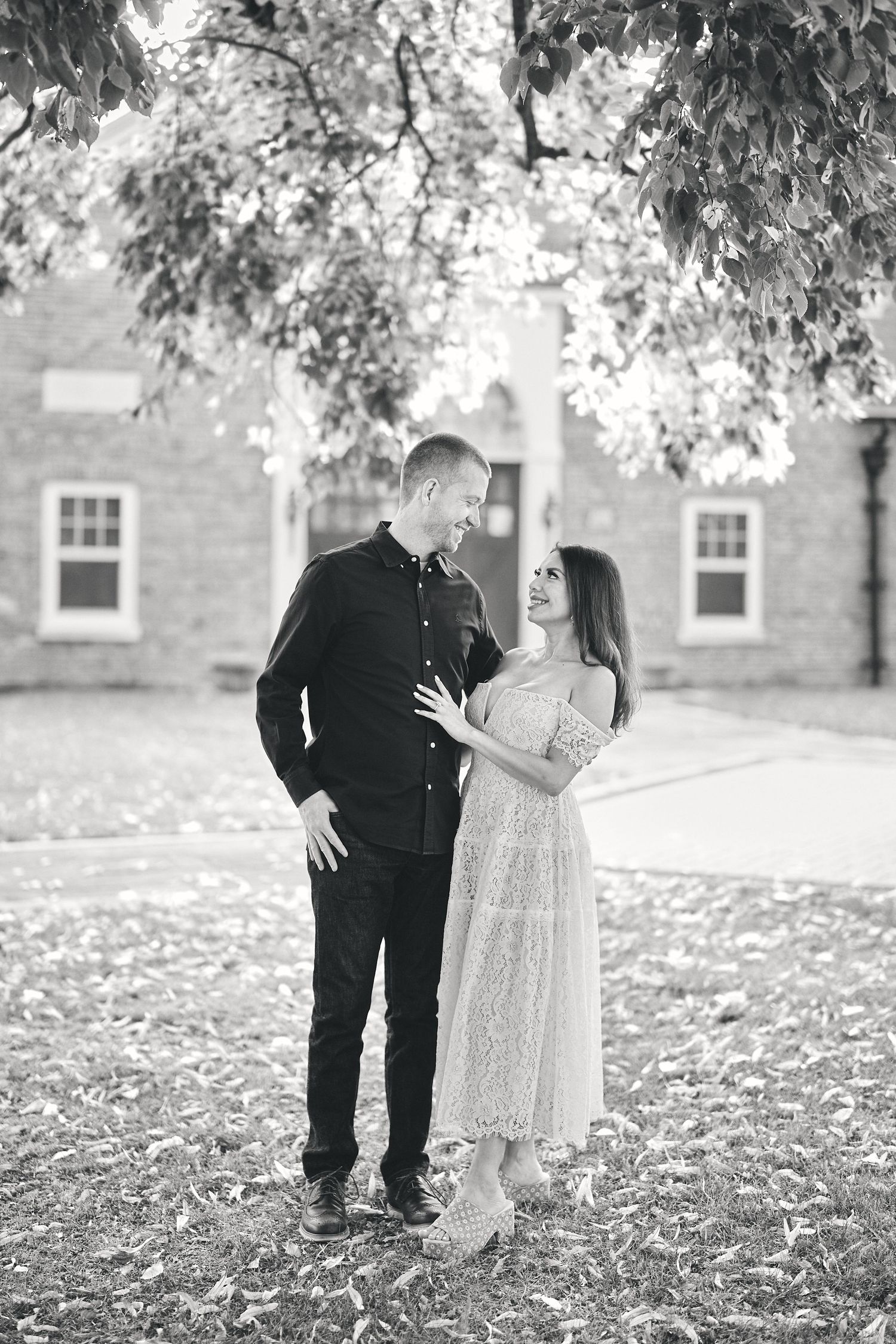 A romantic black and white portrait of a smartly dressed couple standing close together under autumn trees.