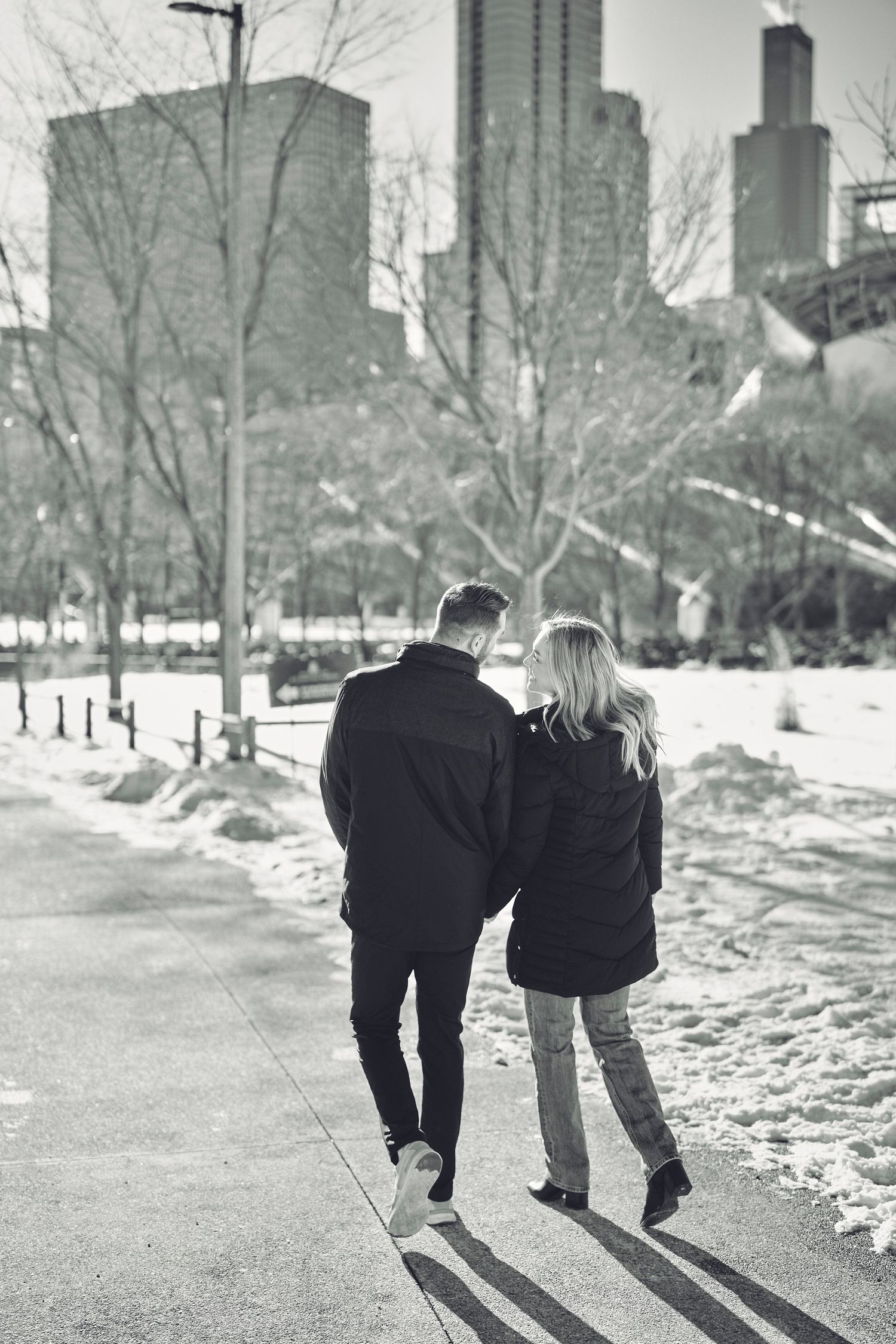 A couple walks together on a snowy path in a city park with tall buildings in the background on a winter day.