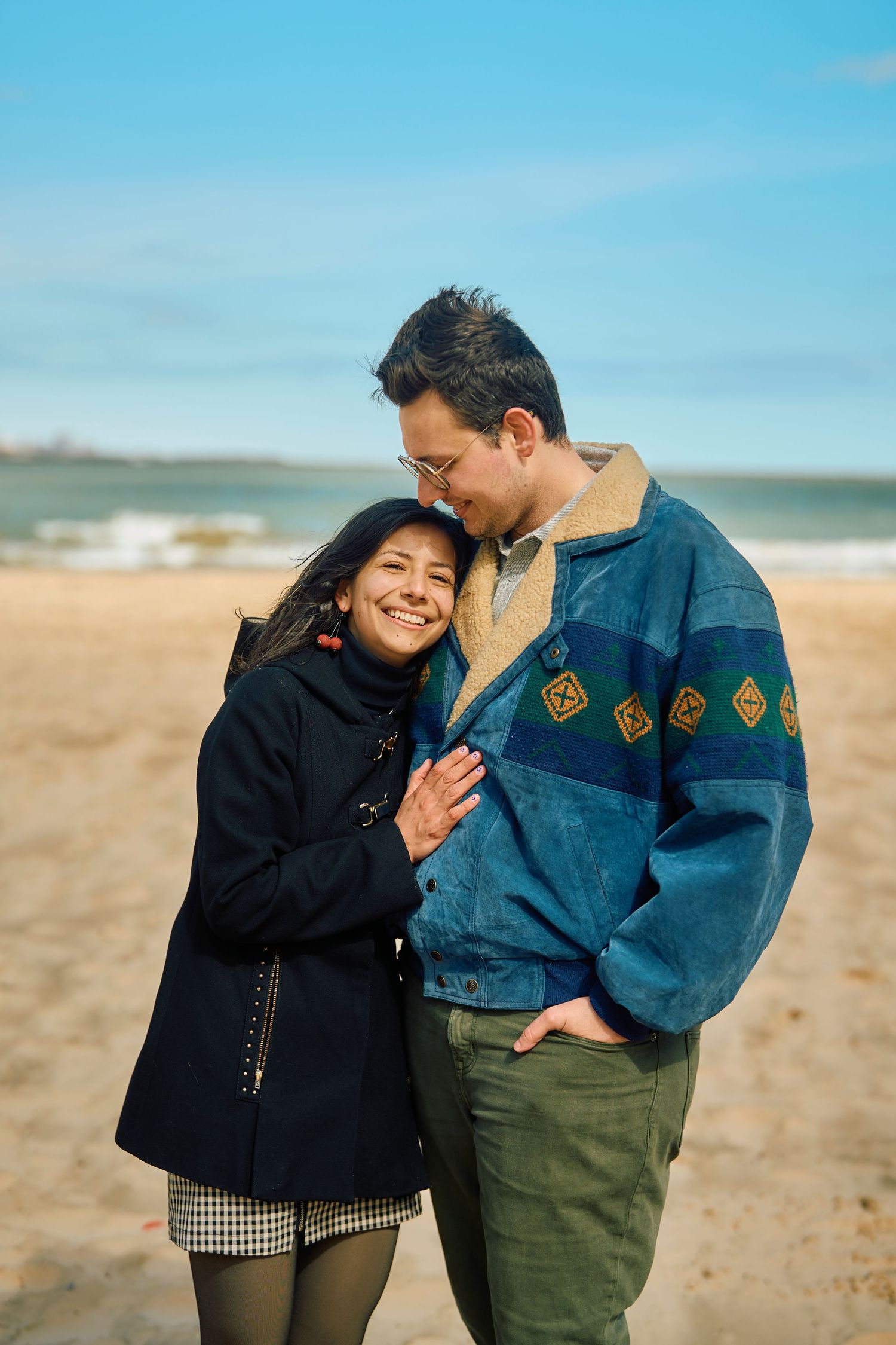 A couple embraces and smiles together on a sandy beach with blue sky and ocean in the background.