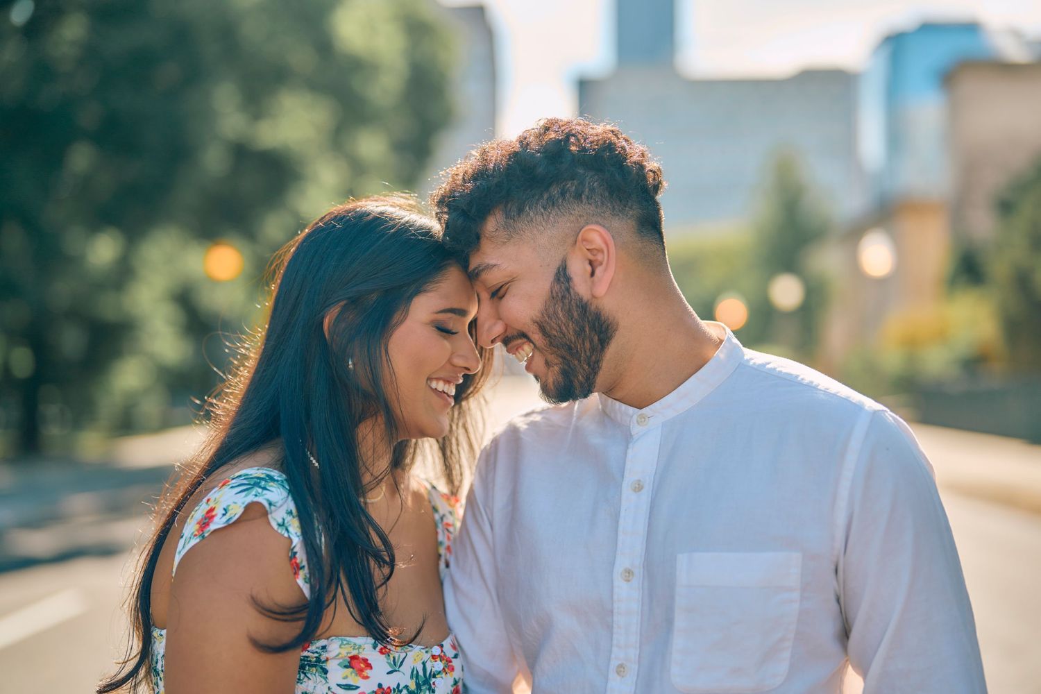 A couple shares an intimate moment together on a sunny day with an urban backdrop and soft natural lighting.