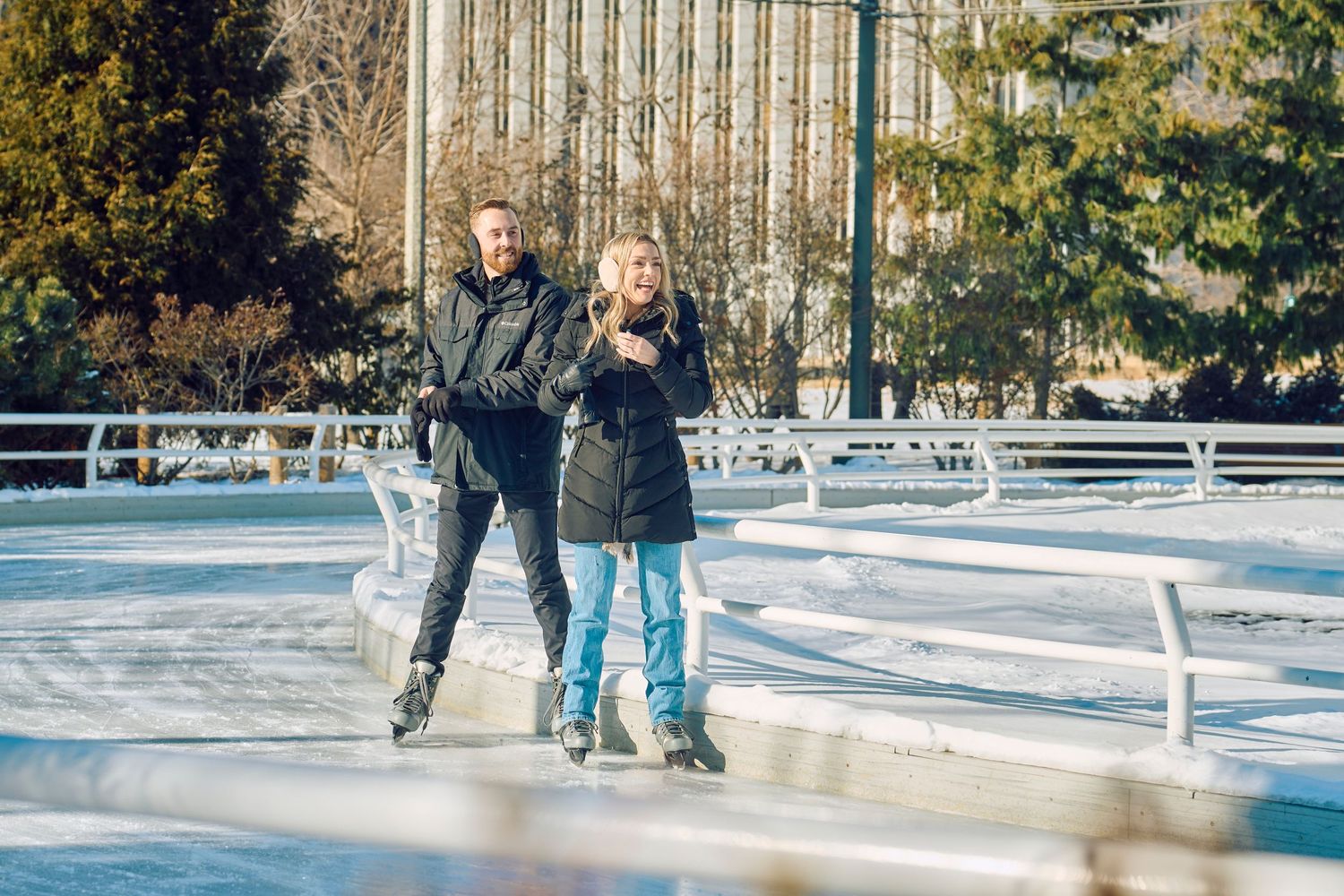 Two people ice skating together on an outdoor rink surrounded by buildings and trees on a sunny winter day.