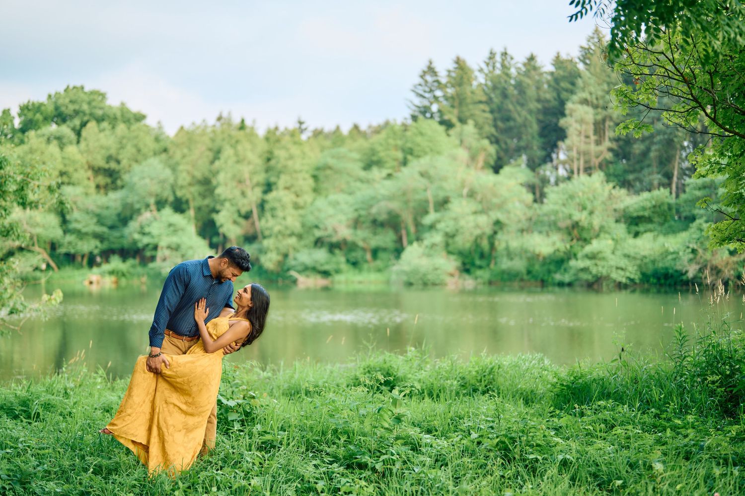 A romantic couple shares an intimate moment by a serene lake surrounded by lush green trees.
