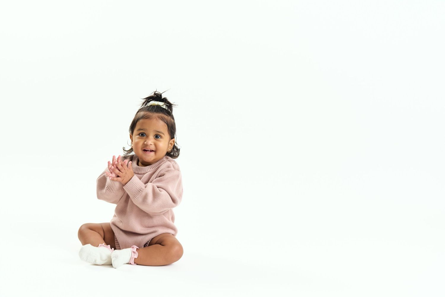 A baby in a pink sweater sits and smiles while playing in a white studio setting.