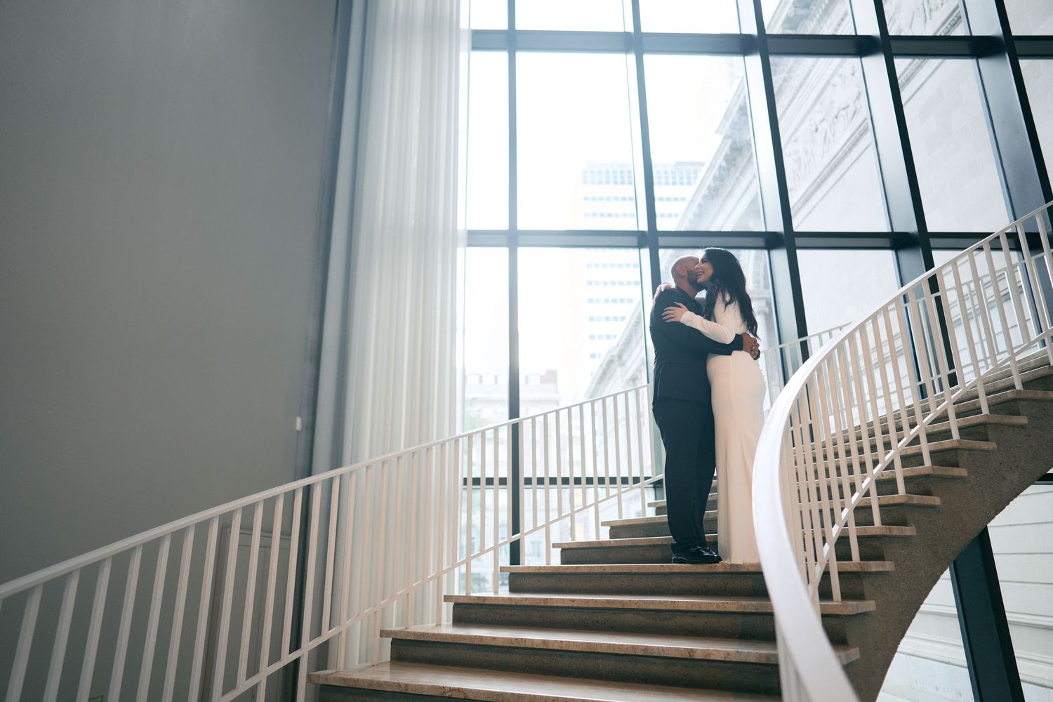 A romantic couple sharing a tender moment on an elegant curved staircase with white railings and natural light streaming in.