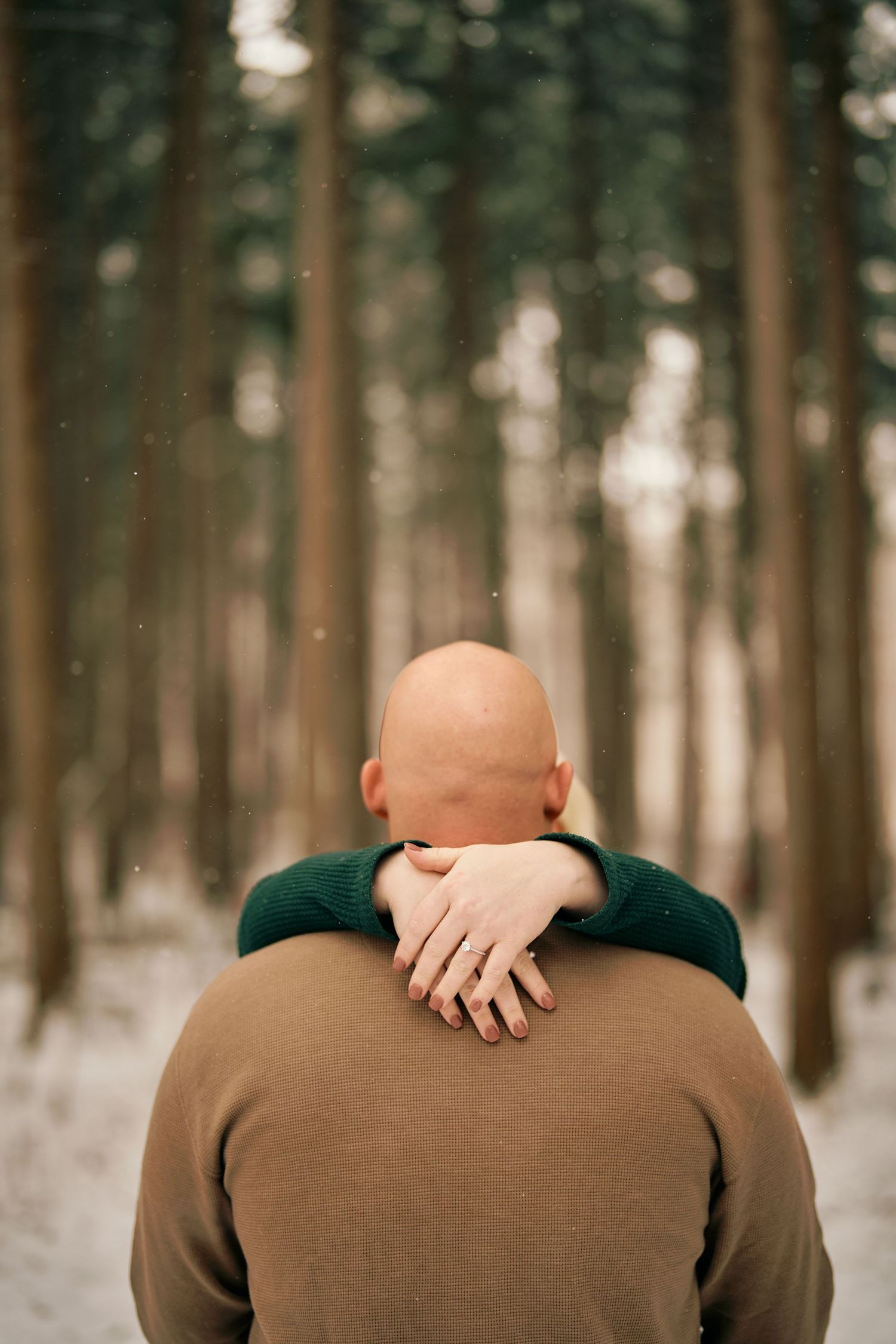 View from behind of someone in brown sweater hugging another person in green in a snowy forest setting.
