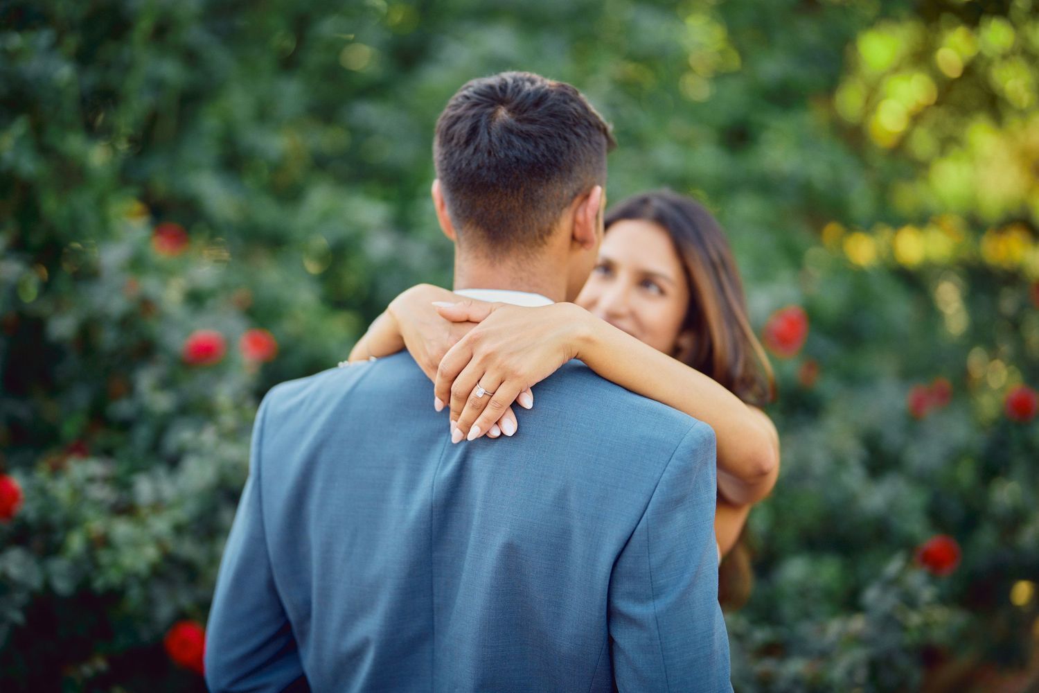 A couple embraces in a garden setting with blurred red flowers and greenery in the background.