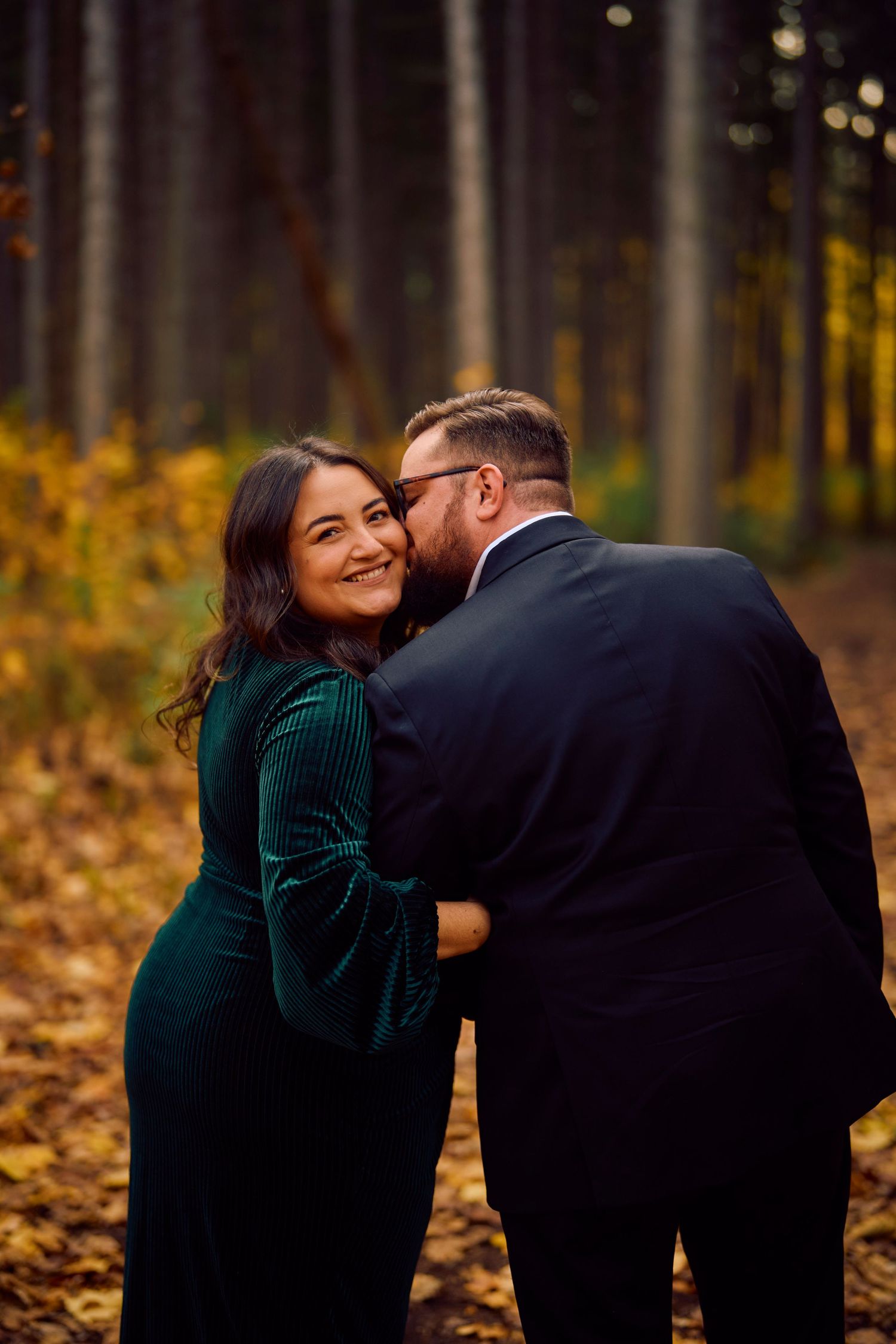 A couple sharing an intimate moment in a forest with autumn leaves on the ground.