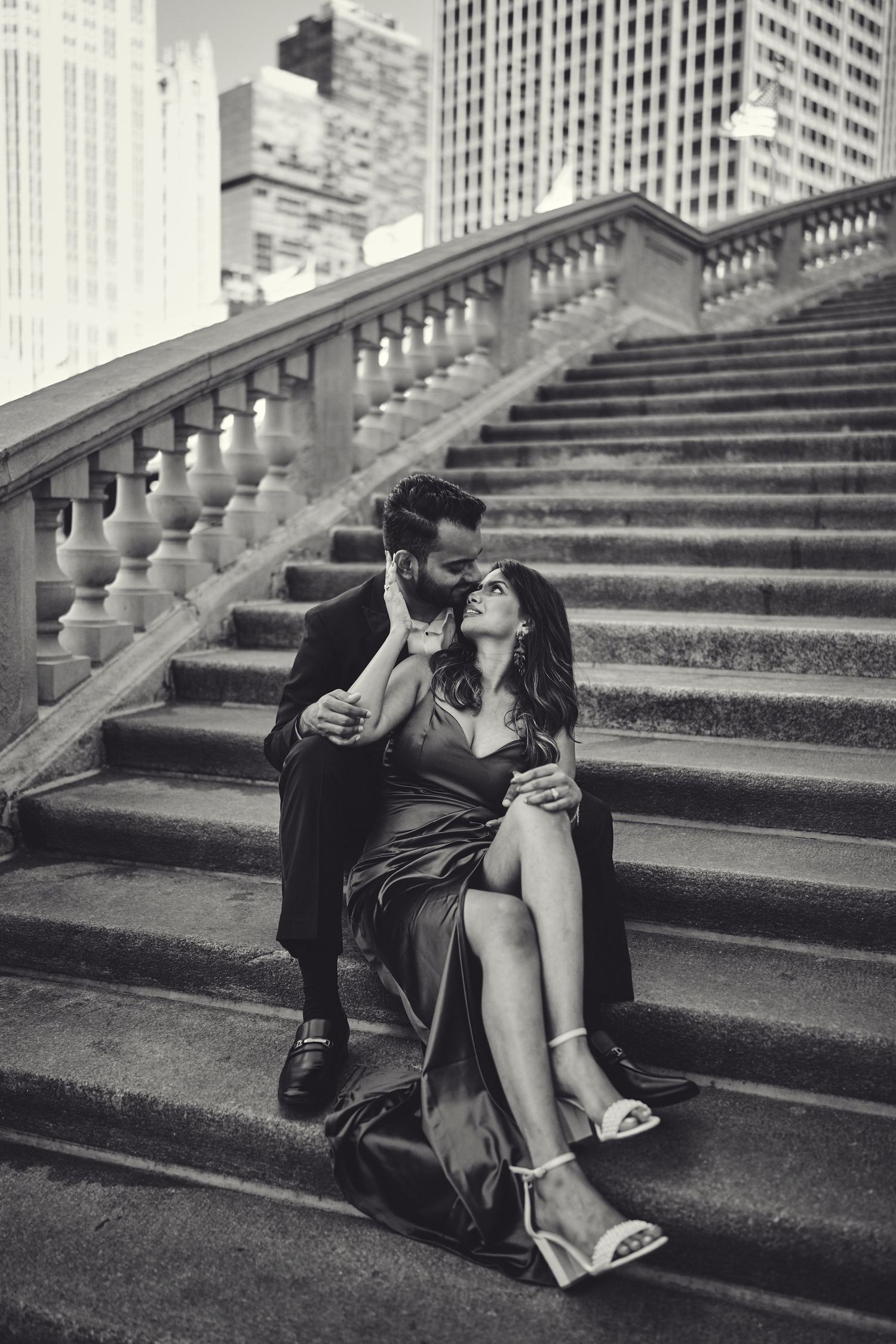 A romantic black and white urban photograph of a couple embracing on elegant stone steps with city buildings in background.