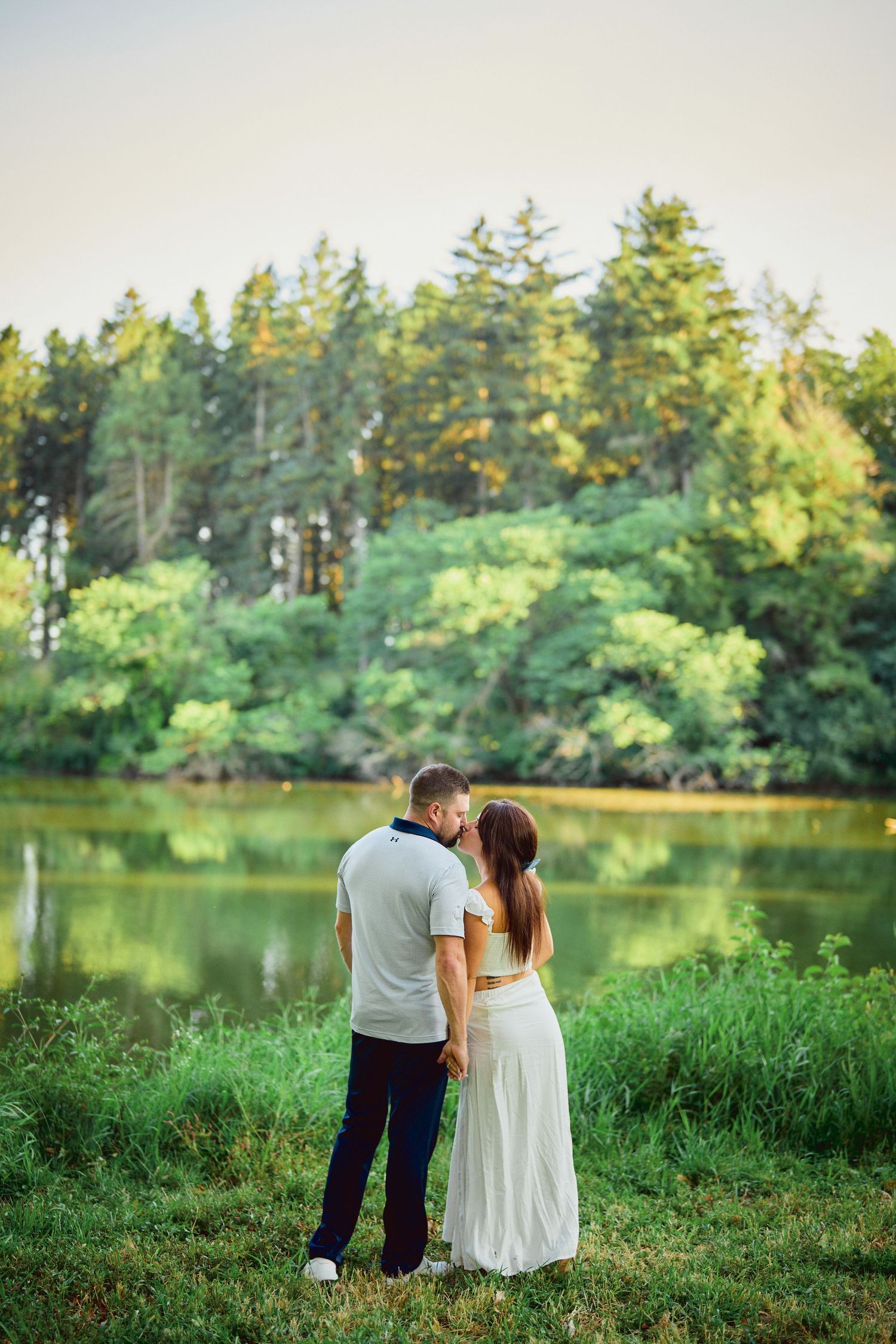 A couple stands together by a serene lake surrounded by evergreen trees and lush greenery on a summer day.