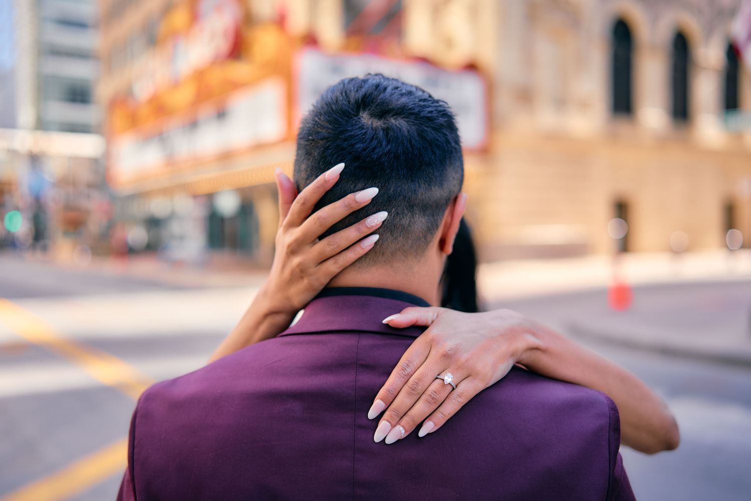 Person wearing a purple shirt embraces someone on a city street, showing long acrylic nails on their hand.
