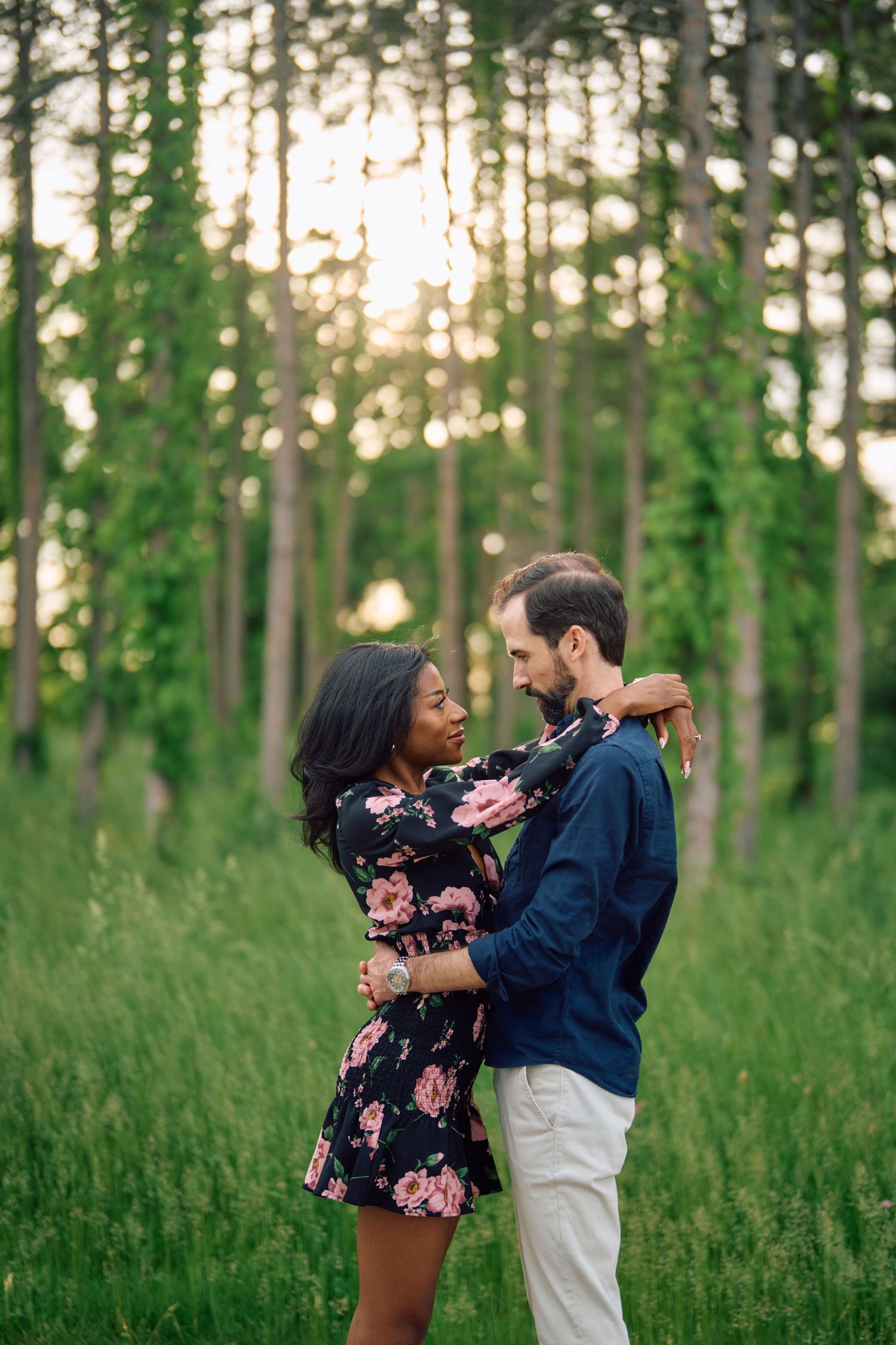 A romantic couple embraces in a sunlit forest meadow, with her wearing a floral dress and him in a navy shirt.