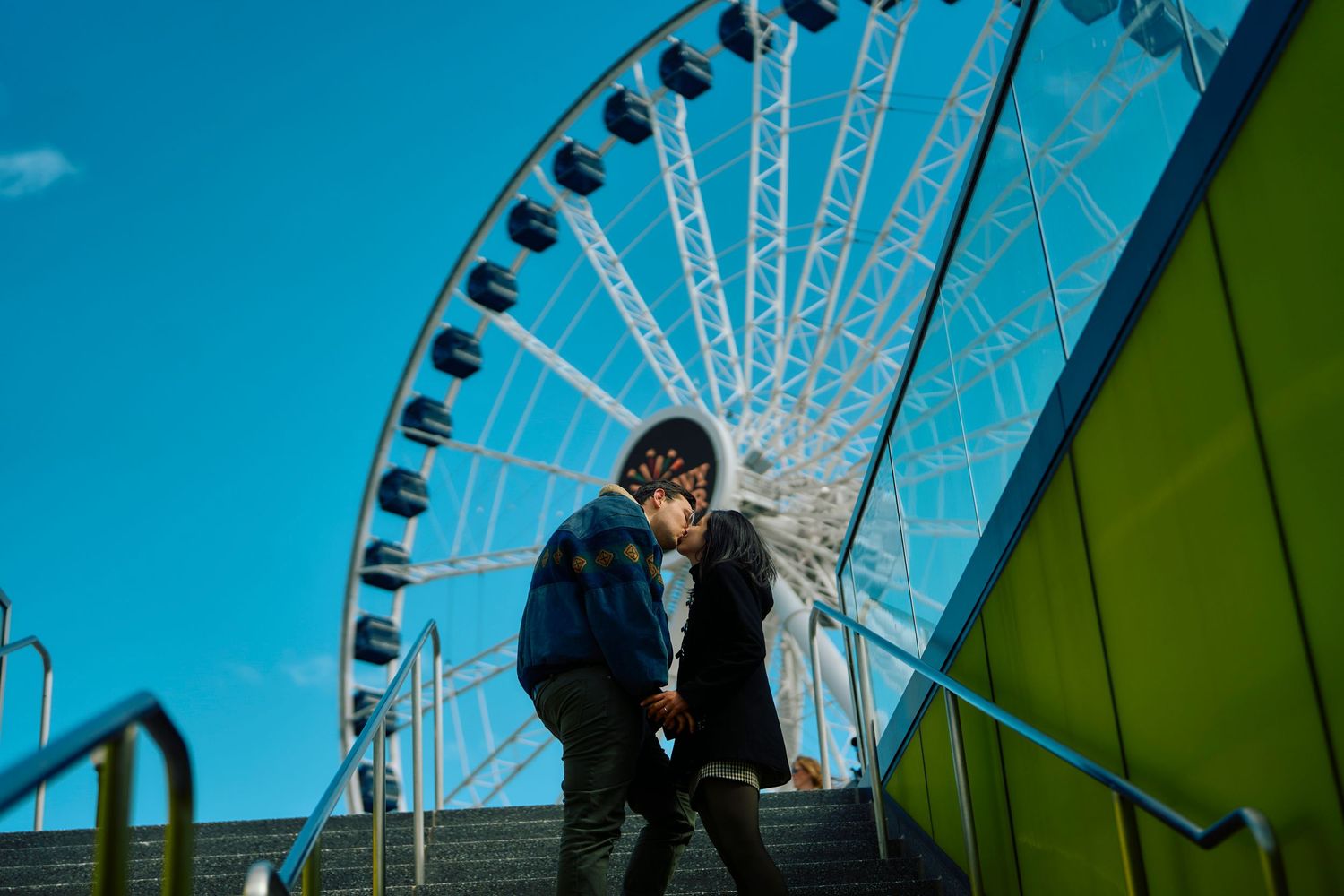 A person in dark clothing sits on steps with a towering white ferris wheel against bright blue sky in the background.