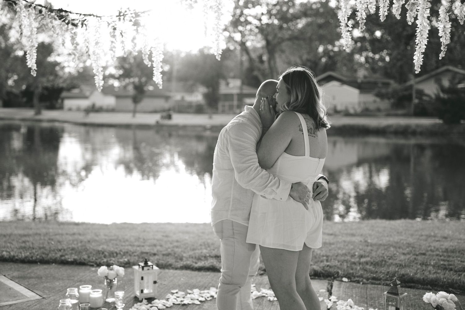 A romantic black and white photo of a couple embracing by a serene lake at sunset with flowers on the ground.