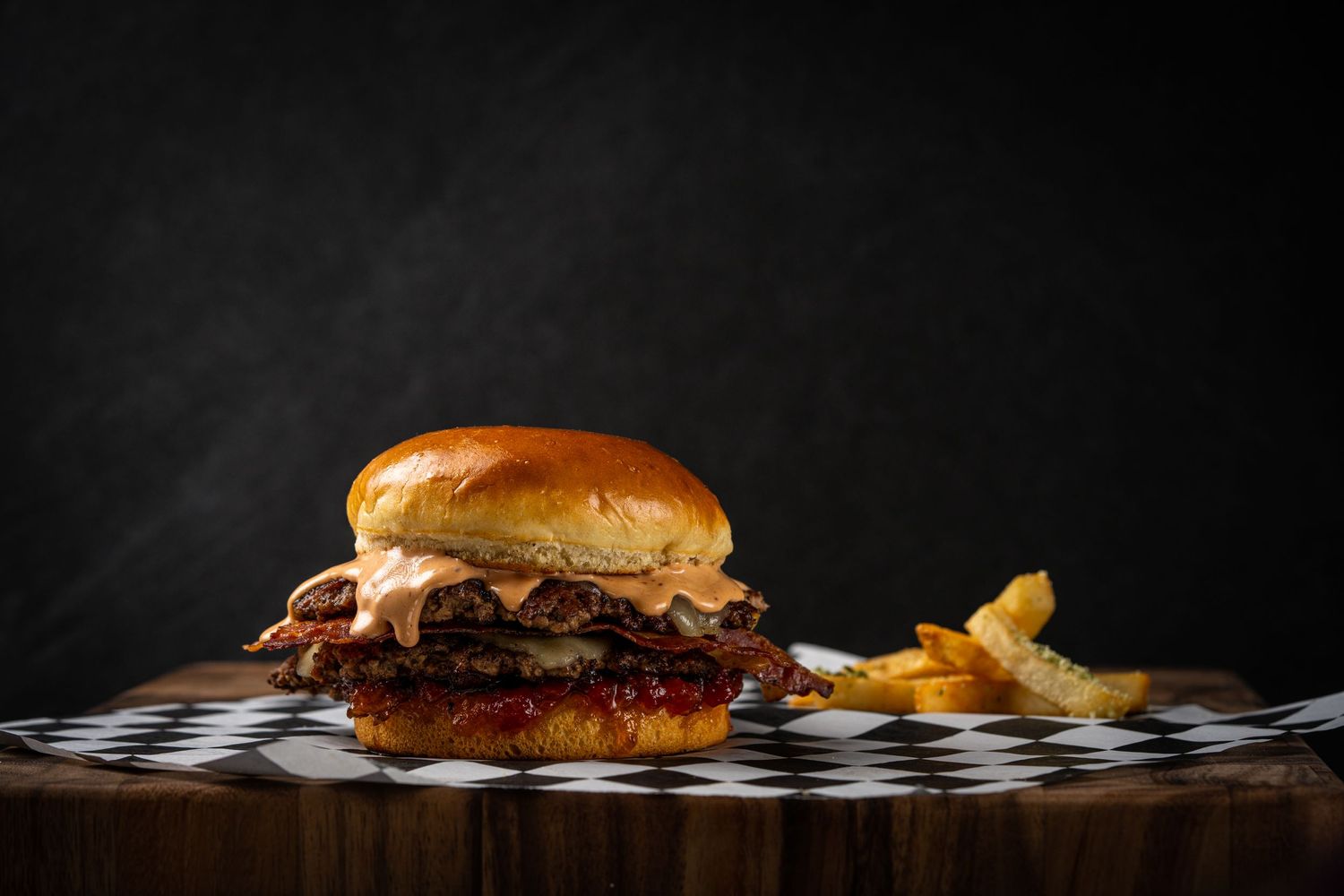 Gourmet burger with fries served on checkered paper on wooden surface against dark background.